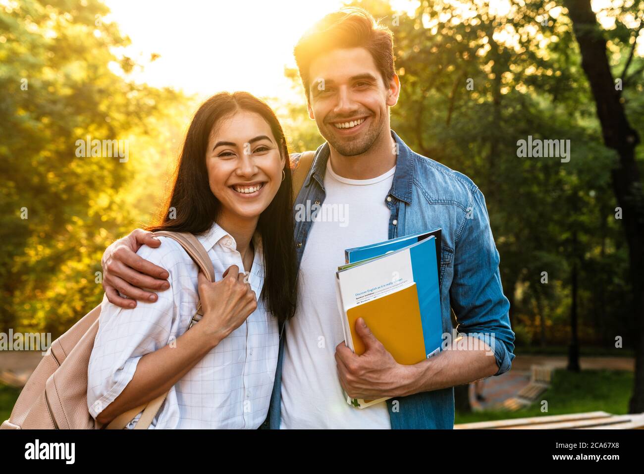 Bild eines fröhlichen multikulturellen Studentenpaares umarmt und lächelt beim Spaziergang im grünen Park Stockfoto