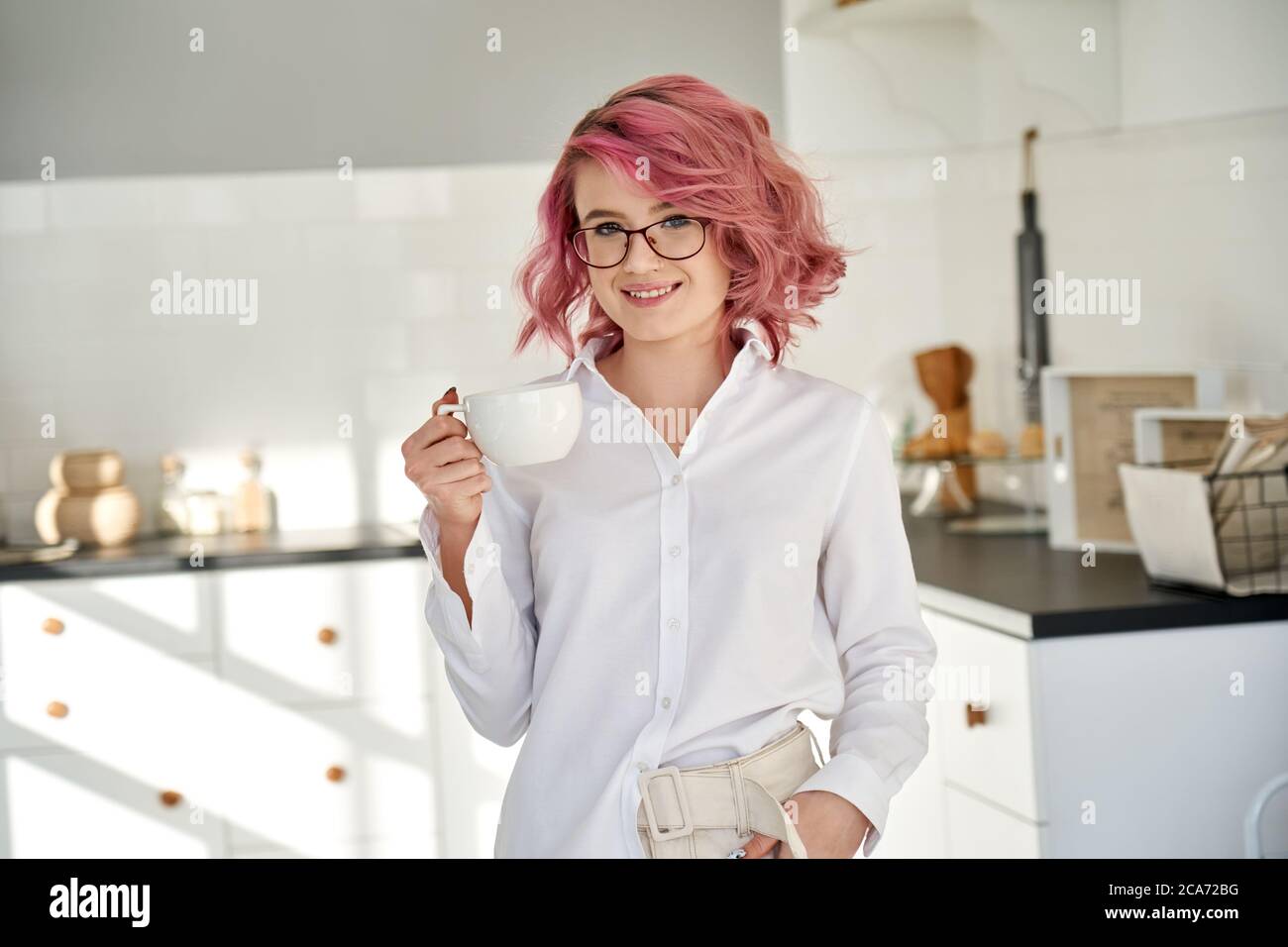 Lächelndes junges Teenager-Mädchen trägt weißes Hemd und Gläser trinken Tee in der Küche. Stockfoto