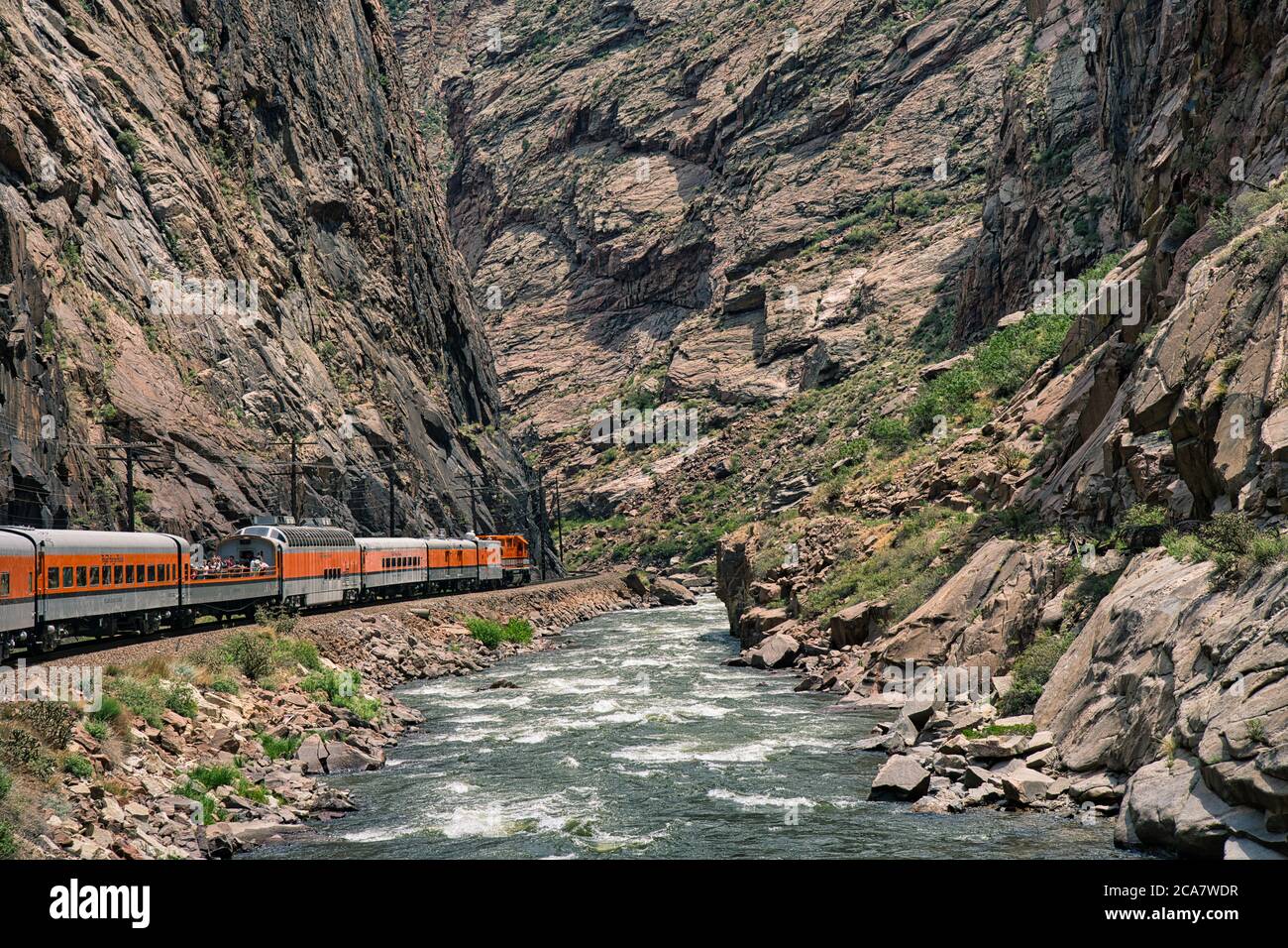 Trainieren Sie tief in der königlichen Schlucht neben dem arkansas Fluss in Colorado. Orange Motor neben rauschendem blauem Wasser mit felsigen Klippen ringsum Stockfoto