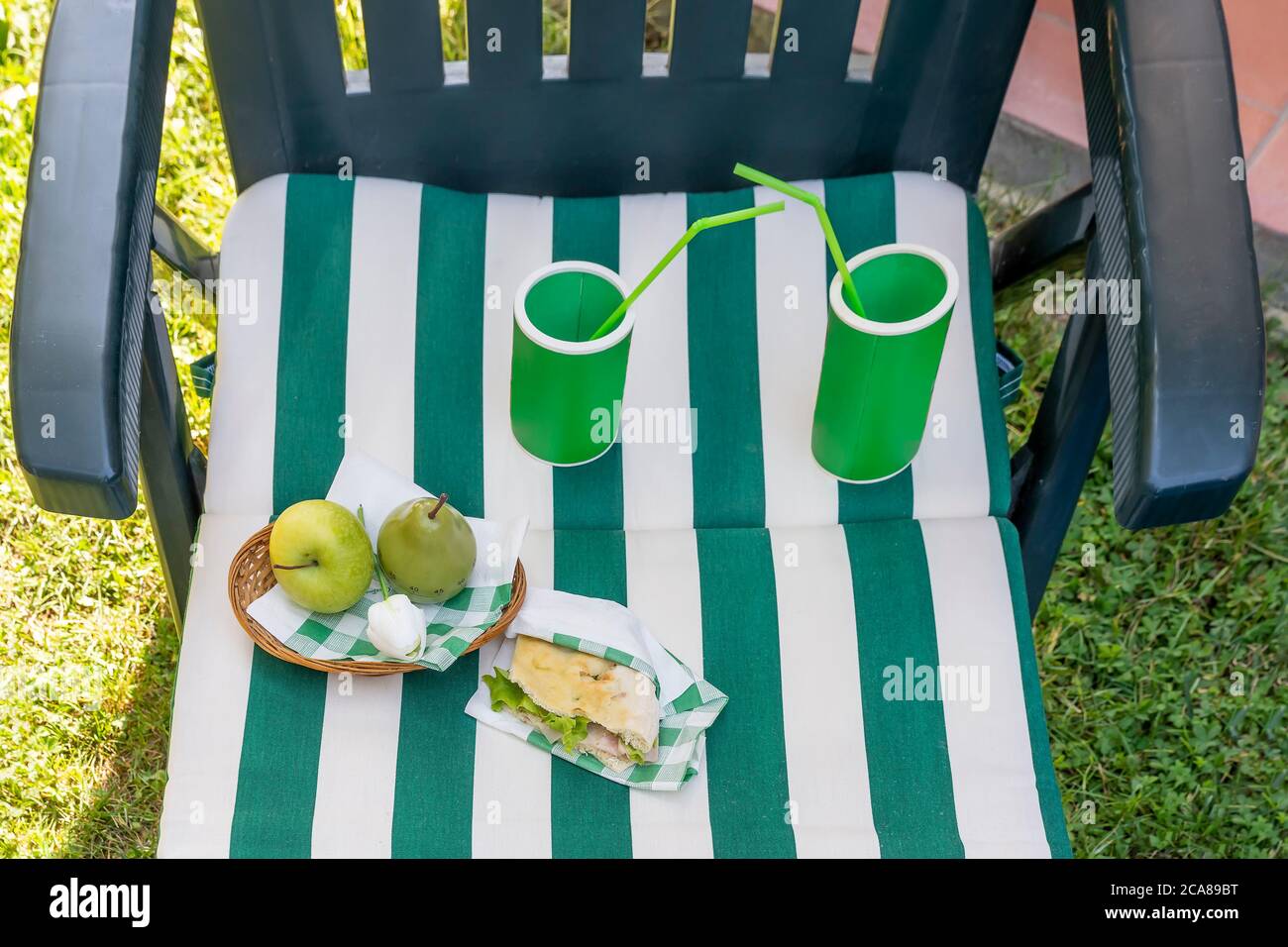 Picknick-Komposition in grünen Sonnenbetten mit weiß-grün gestreiften Matratzen, Kühler mit Strohhalmen, Sandwich und Obst Stockfoto