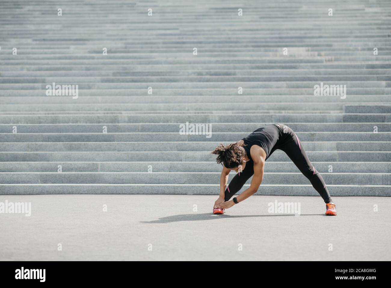 Dehnung nach dem Training. Afroamerikanische Frau mit Fitness-Tracker und kabellosen Kopfhörern macht Übungen für Beine Stockfoto