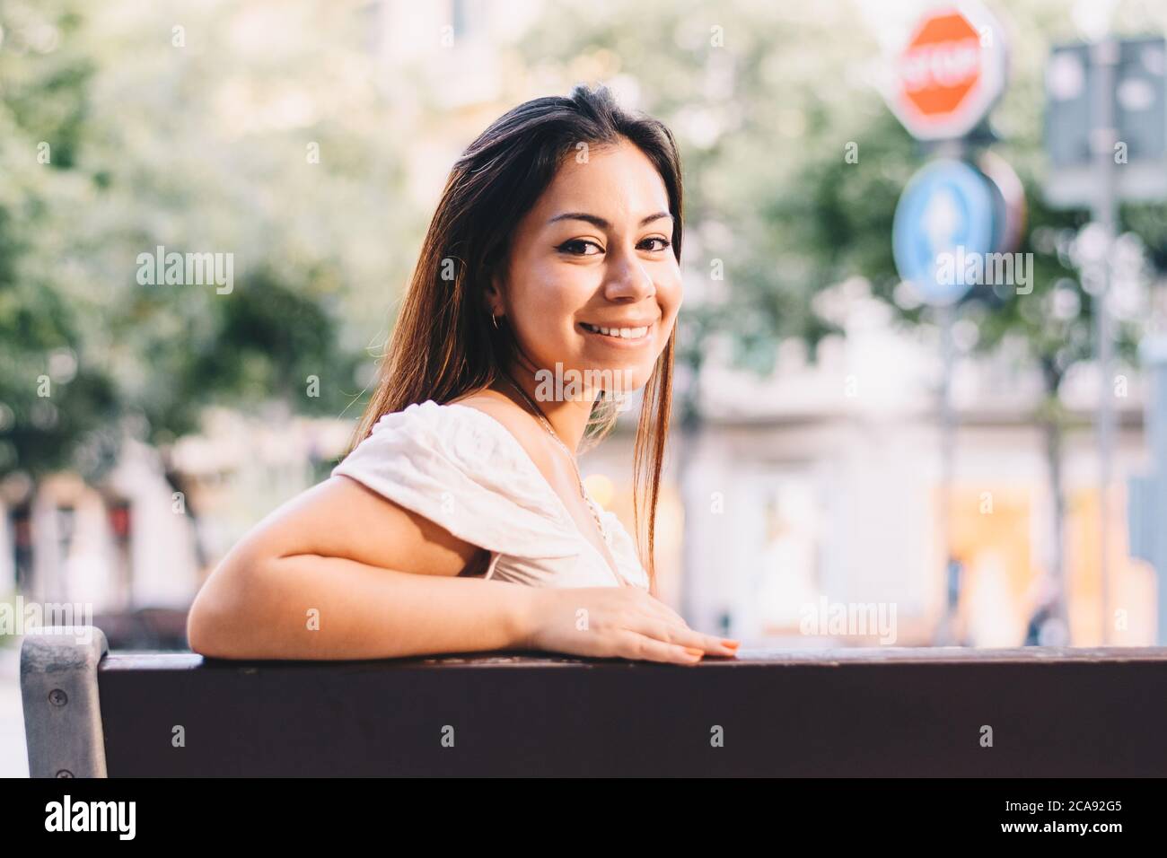 Eine Latina-Frau, die auf einer Stadtbank sitzt und die Kamera anlächelt Stockfoto