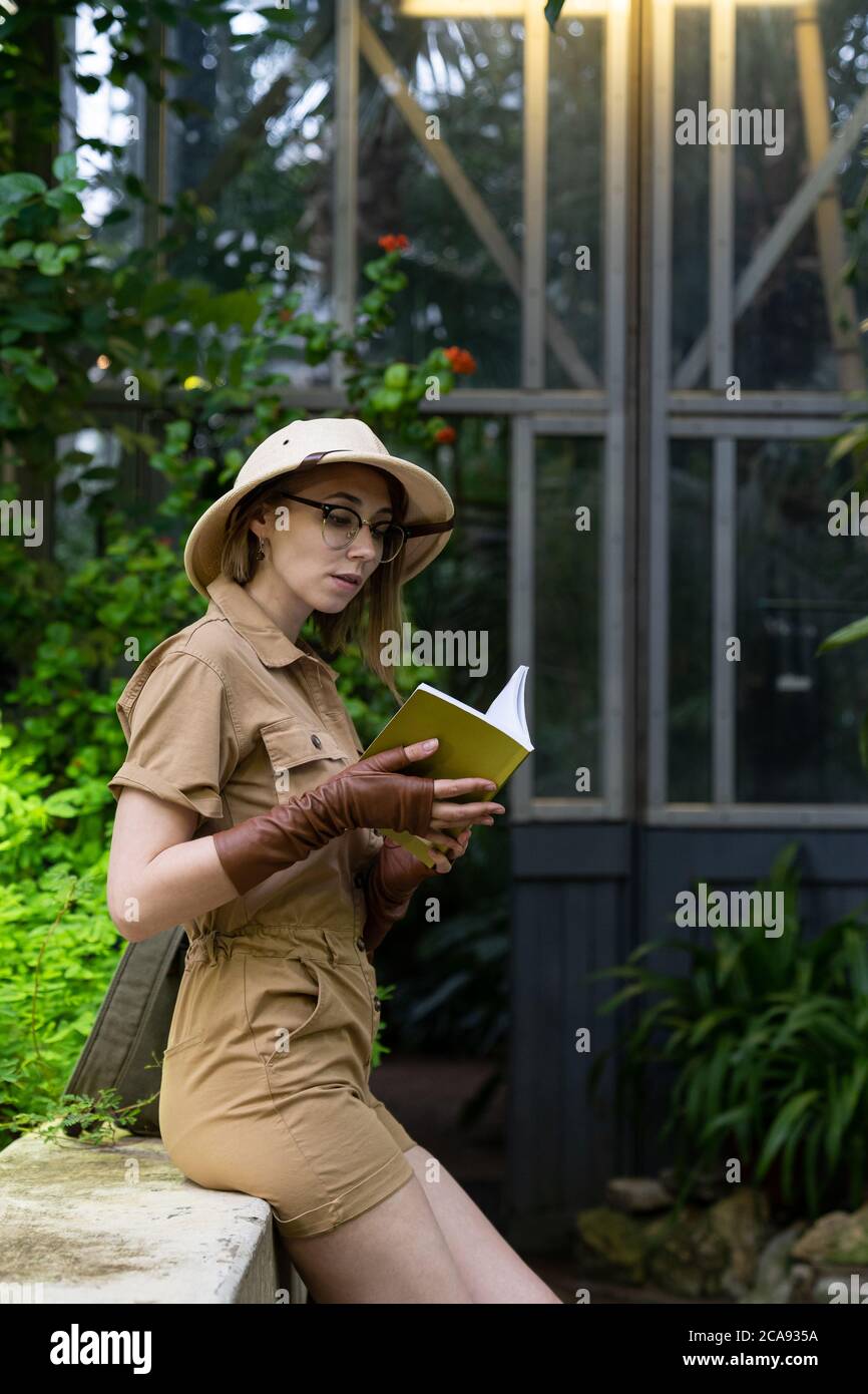 Frau Botanikerin im Safaristil im Gewächshaus gekleidet. Naturforscher trägt Hut und khaki Kleidung, leaver Handschuhe ruhen und lesen ein Buch umgeben von Stockfoto