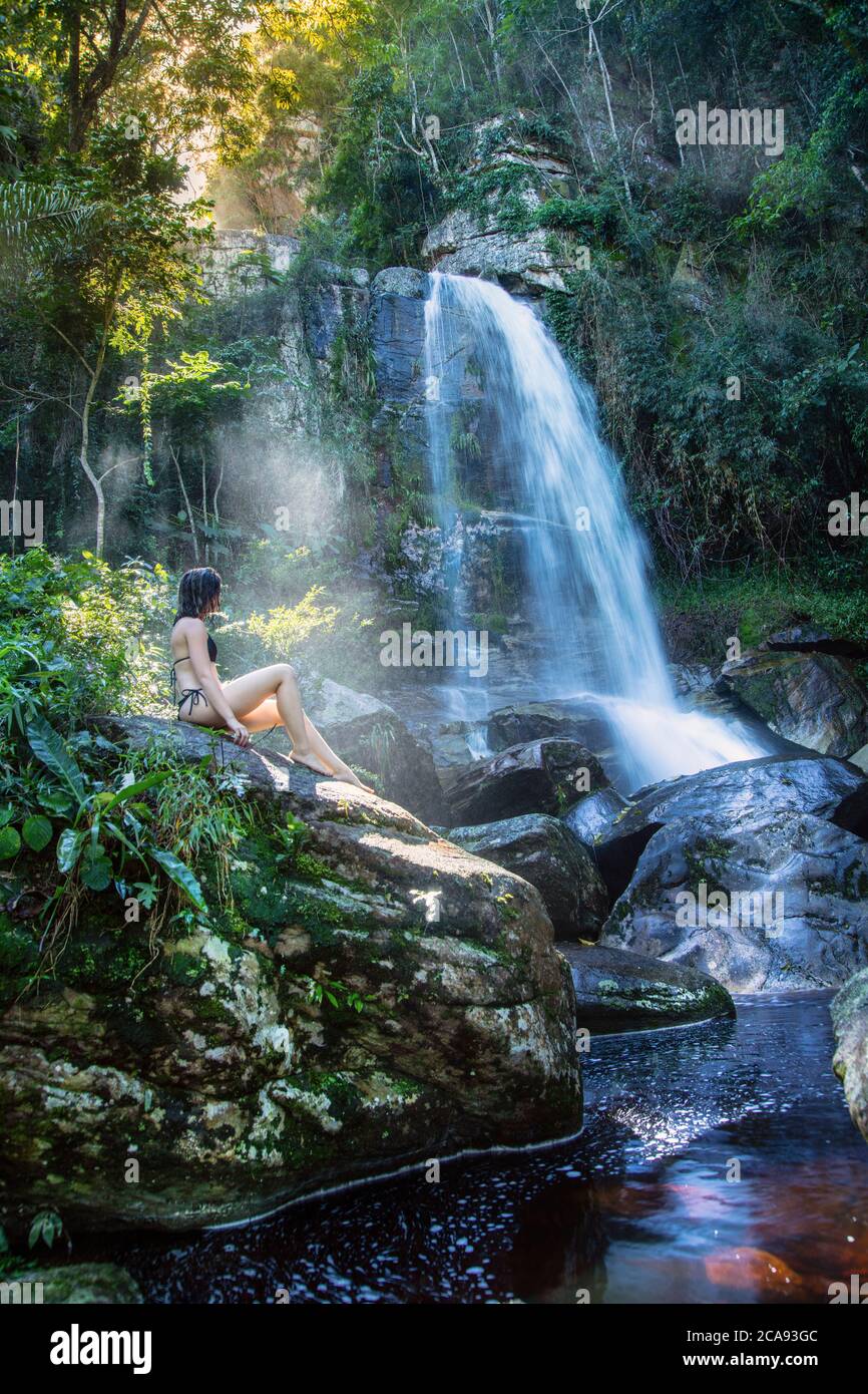 Eine schöne junge Frau, die in einem Sonnenstrahl vor einem unberührten Wasserfall im Regenwald, Brasilien, Südamerika sitzt Stockfoto