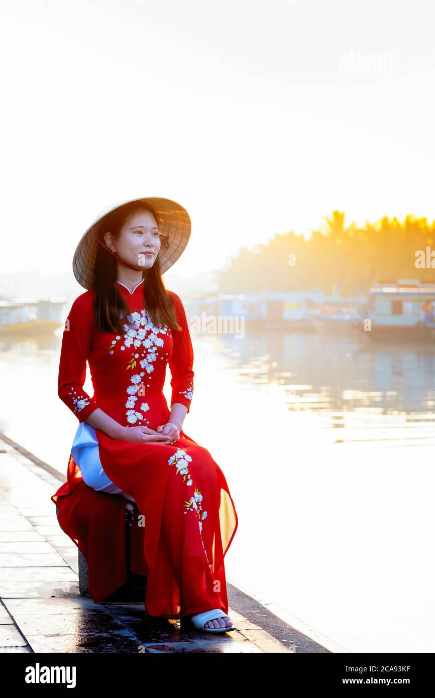 Eine junge asiatische Frau in einem roten Ao Dai Kleid und konischen Hut lächelnd und sitzt draußen neben einem Fluss in Hoi an, Vietnam, Indochina Stockfoto