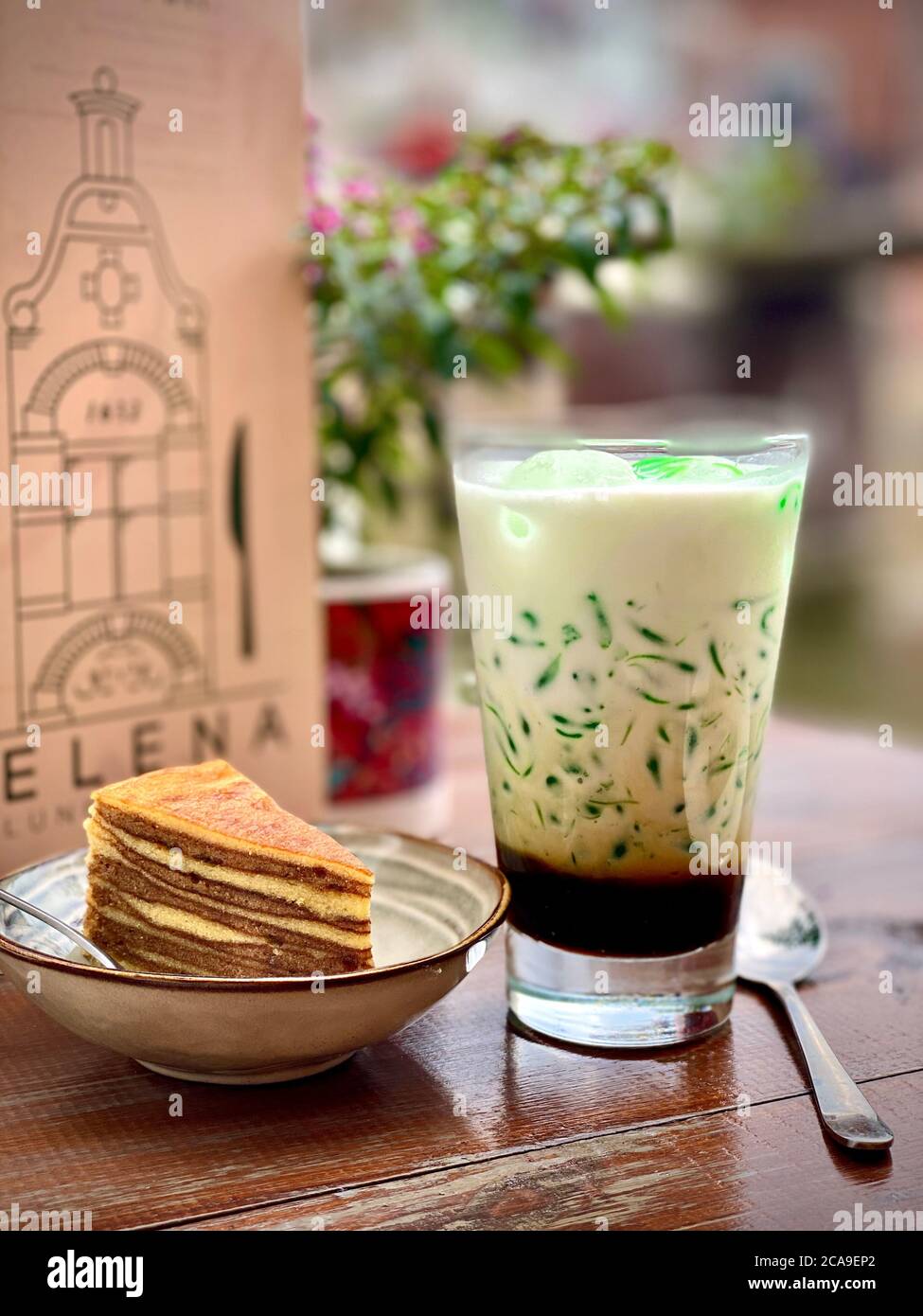 Traditionelles indonesisches süßes Erfrischungsgetränk Cendol aus Pandas-Saft, Mungbohne. Restaurant Cafe Holztisch draußen. Serviertes Getränk im Glas mit einem Stück Schichtkuchen in einer Schüssel mit einem Menü im Hintergrund. Bunt grün, braun cremig Getränk. Durstlöschender Durst. Stockfoto