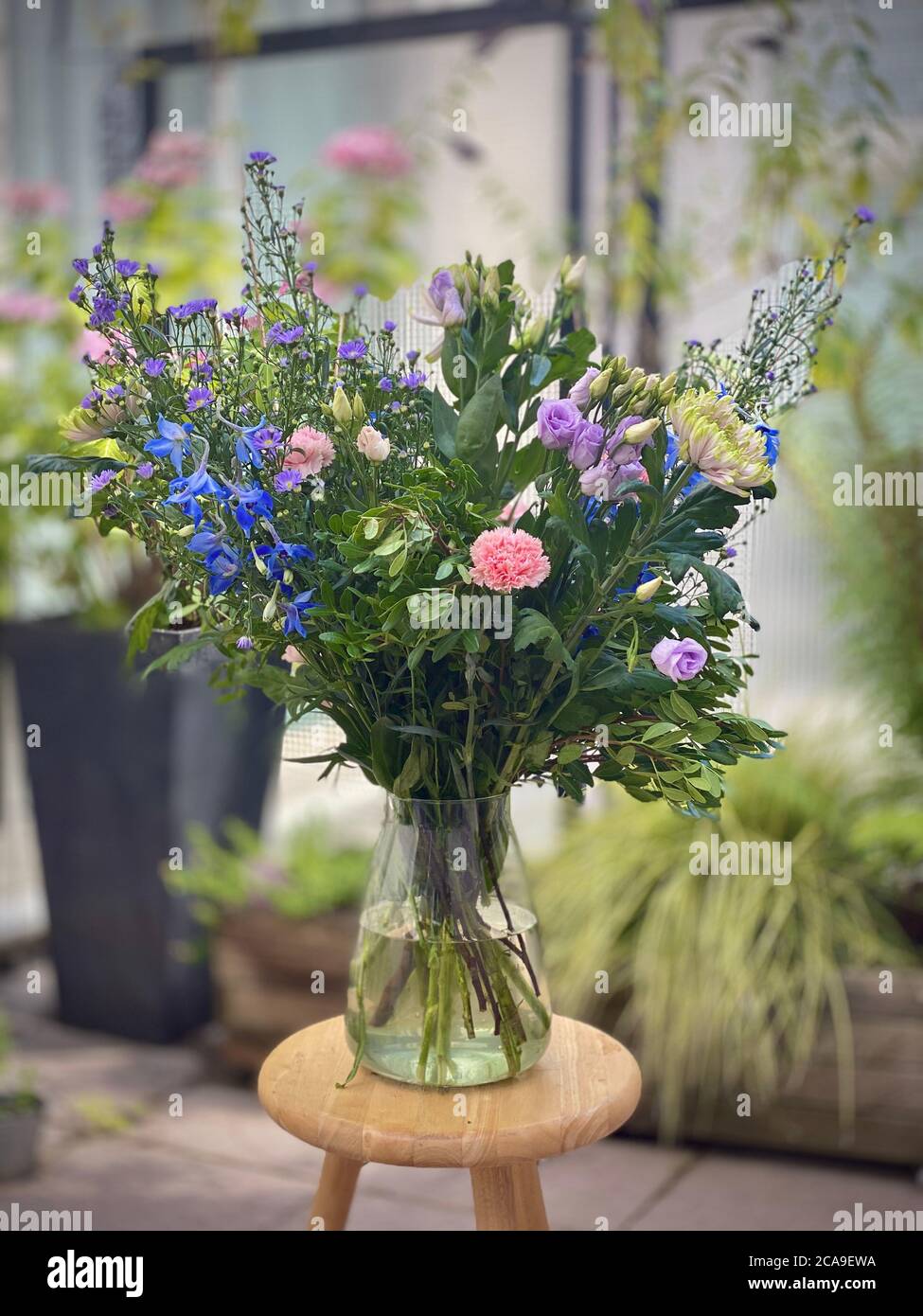 Schöne große Blumenstrauß in einer Vase draußen auf einem kleinen Holztisch angezeigt. Plantlife blühende im Wasser stehend. Verschwommener Hintergrund, blau-lila und weiße Nelken grün-blättrige Blätter. Stockfoto