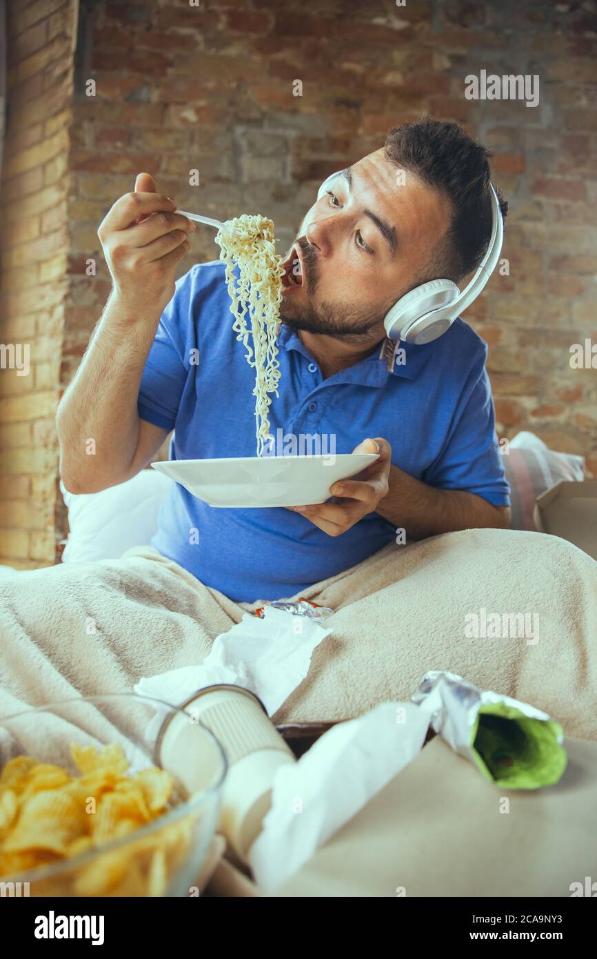 Leidenschaftlich essen Instant Nudel. Fauler Mann, der in seinem Bett lebt, umgeben von Unordnung. Man muss nicht ausgehen, um glücklich zu sein. Mit Gadgets, Film und Serien, sieht emotional. Essen Snacks und Fast Food. Stockfoto