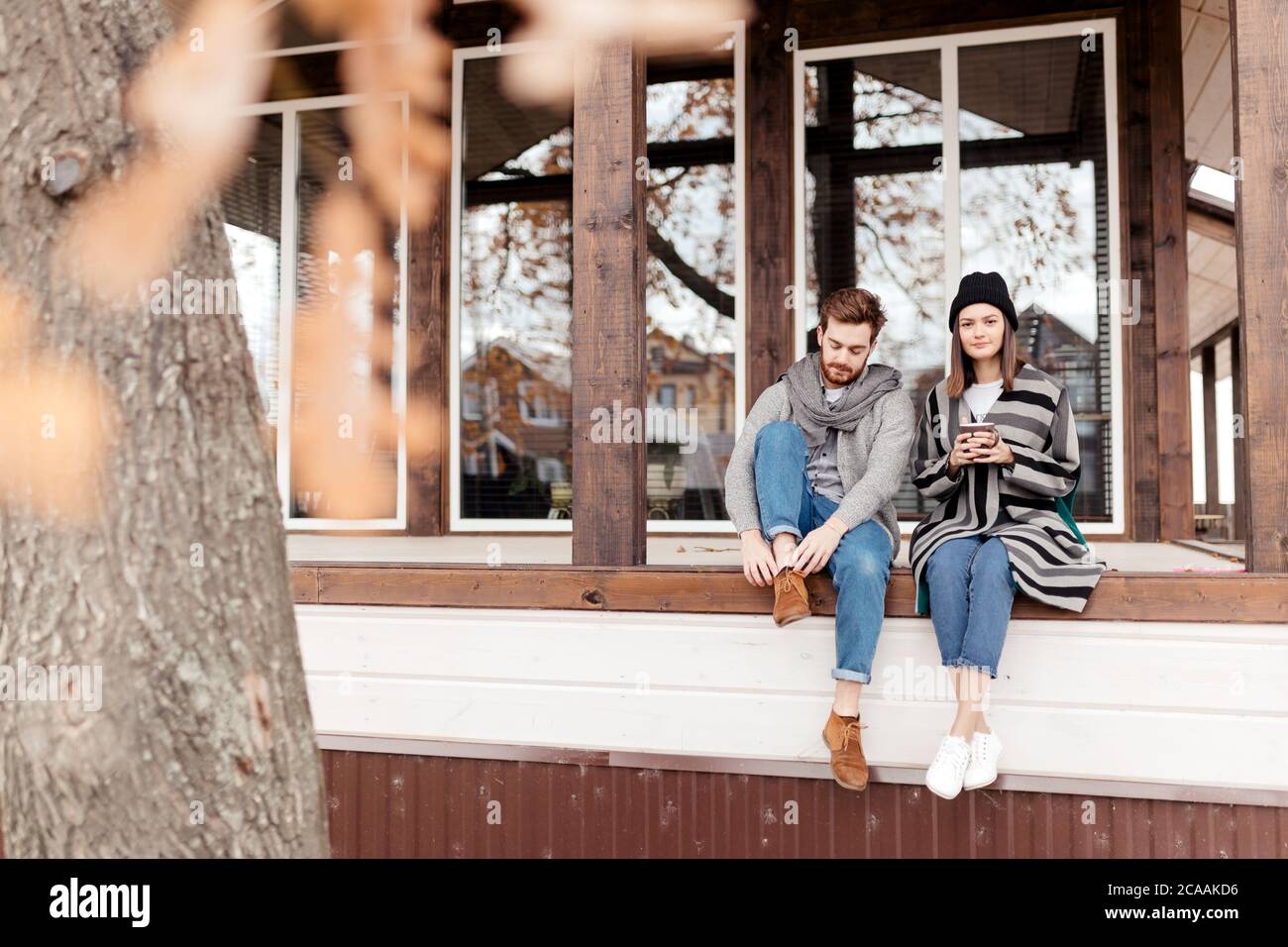 Junges Paar in stilvolle warme Mäntel sitzen mit Schalen auf der Terrasse des modernen Hauses genießen Sie einen wunderschönen Herbst im Freien gekleidet anzeigen Stockfoto Junges Paar in stilvolle warme Mäntel sitzen mit Schalen auf der Terrasse des modernen Hauses genießen Sie einen wunderschönen Herbst im Freien gekleidet anzeigen Stockfoto