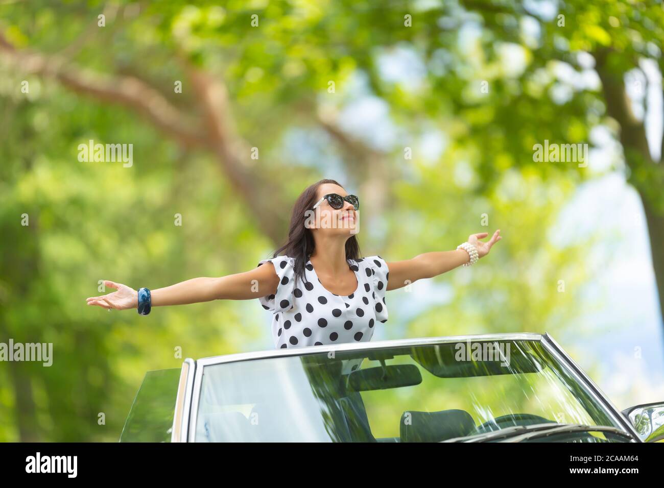 Frau in Sonnenbrille sitzt auf dem Cabrio, Arme weit geöffnet, die Sonne und frische Luft zu saugen. Stockfoto