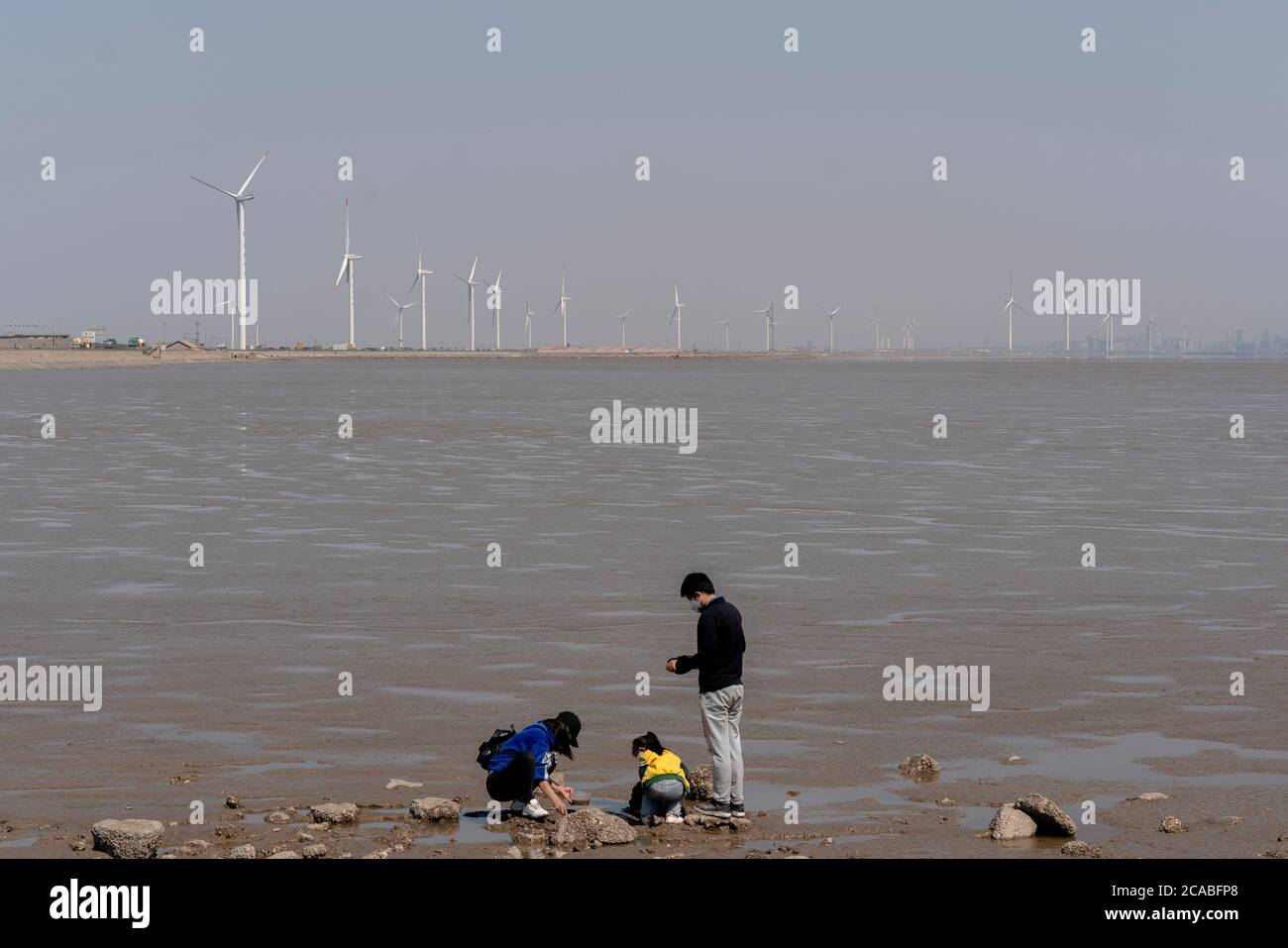 Eine chinesische Familie genießt Freizeit am Meer, am Strand gibt es Reihen von Windkraftanlagen. Stockfoto