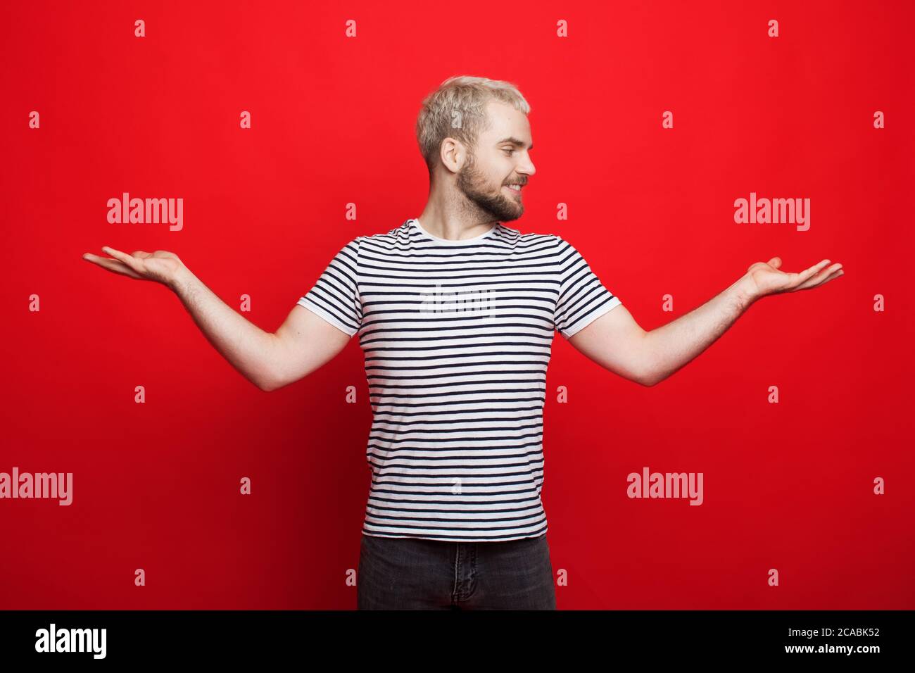 Kaukasischer blonder Mann mit Bart, der zwei Dinge in seinen Handflächen vergleicht, posiert auf einer roten Studiowand Stockfoto