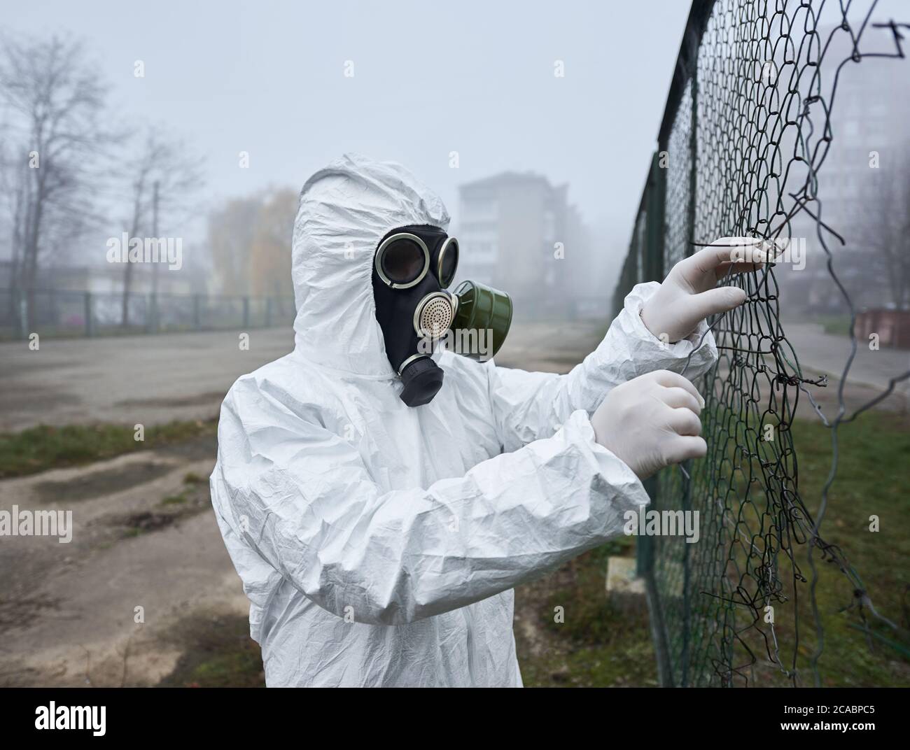 Nahaufnahme eines Wissenschaftlers, der einen zerrissenen Drahtzaun reißt. Mann in Gasmaske und Schutzkostüm, der auf leerem Fußballplatz im Stadthof in der Nähe von Gebäuden steht. Umweltkatastrophe, rauchige Atmosphäre, Nebel. Stockfoto