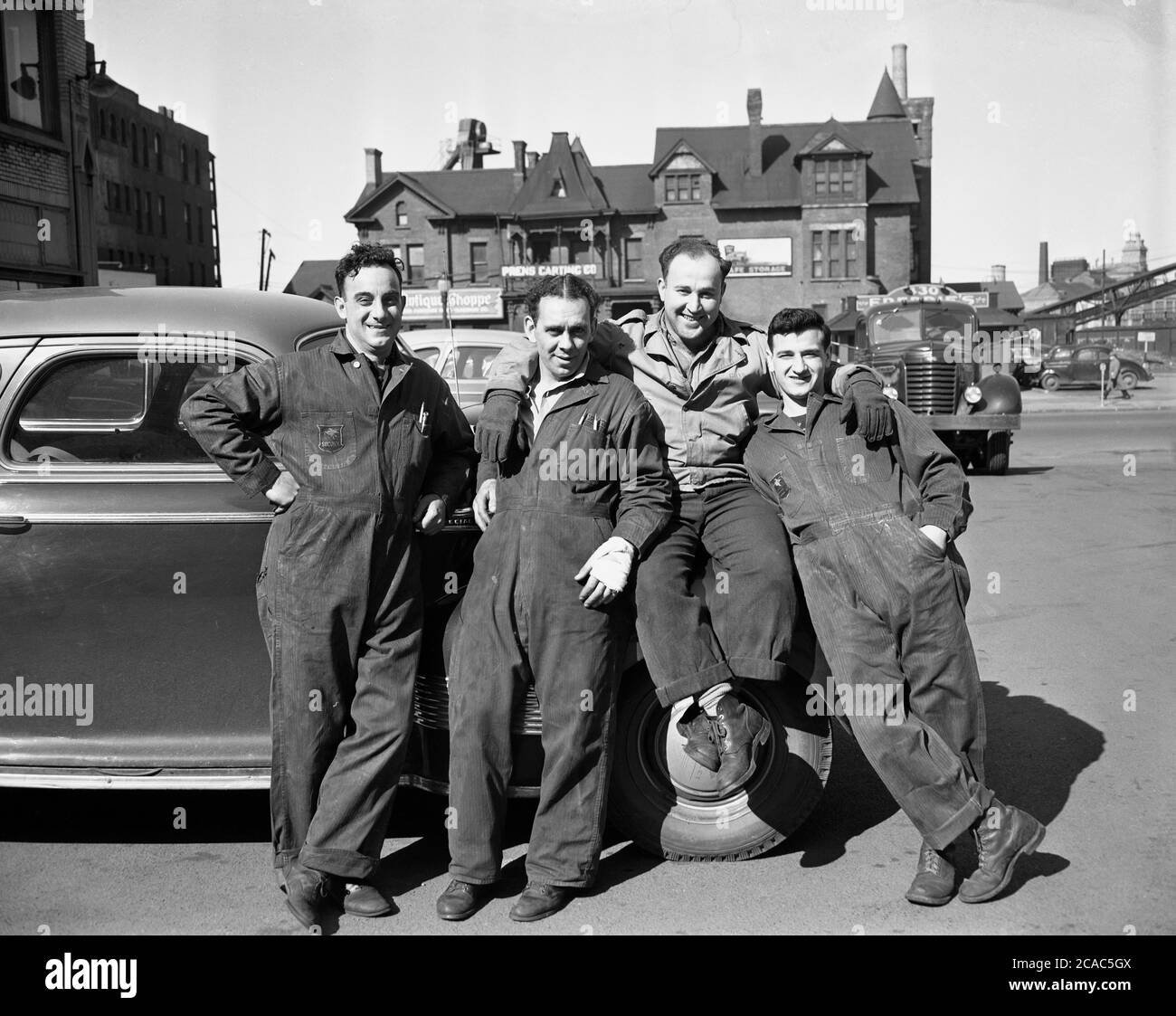 1940er Jahre, historisch, draußen auf dem Platz einer Industriestadt im Mittleren Westen, posieren vier Motormechaniker in ihren Overalls, die sich gegen einen Wagen lehnen, für ein Foto, USA. Ein Schild für die Prens Karting Co ist im Hintergrund zu sehen. Ein Socony-Abzeichen ist auf einem der Overalls des Mechanikers zu sehen, der verkürzte Name für die 'Standard Oil Co of New York'. Stockfoto