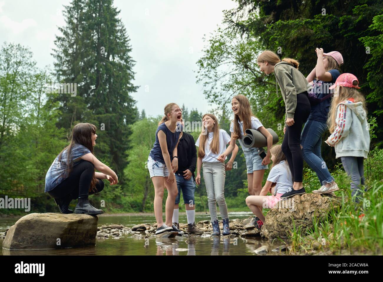 Von unten Blick auf lachende Teens in lässigen Anzügen stehen auf Felsen im Flusswasser im Sommer Tag. Junge Freunde haben Spaß im Freien in der Nähe von grünen Wald. Konzept der Freundschaft, Natur, Camping. Stockfoto