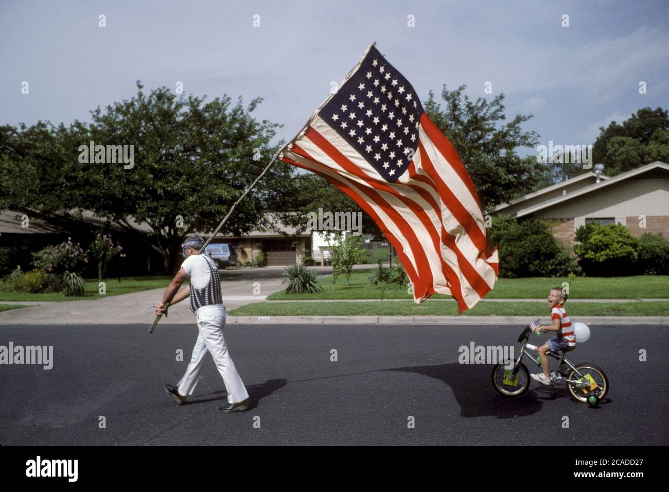 Austin Texas USA: Anglo-Mann mit großer Flagge der Vereinigten Staaten, gefolgt von einem kleinen Jungen auf dem Fahrrad während einer Parade am 4. Juli durch ein Viertel. ©Bob Daemmrich Stockfoto