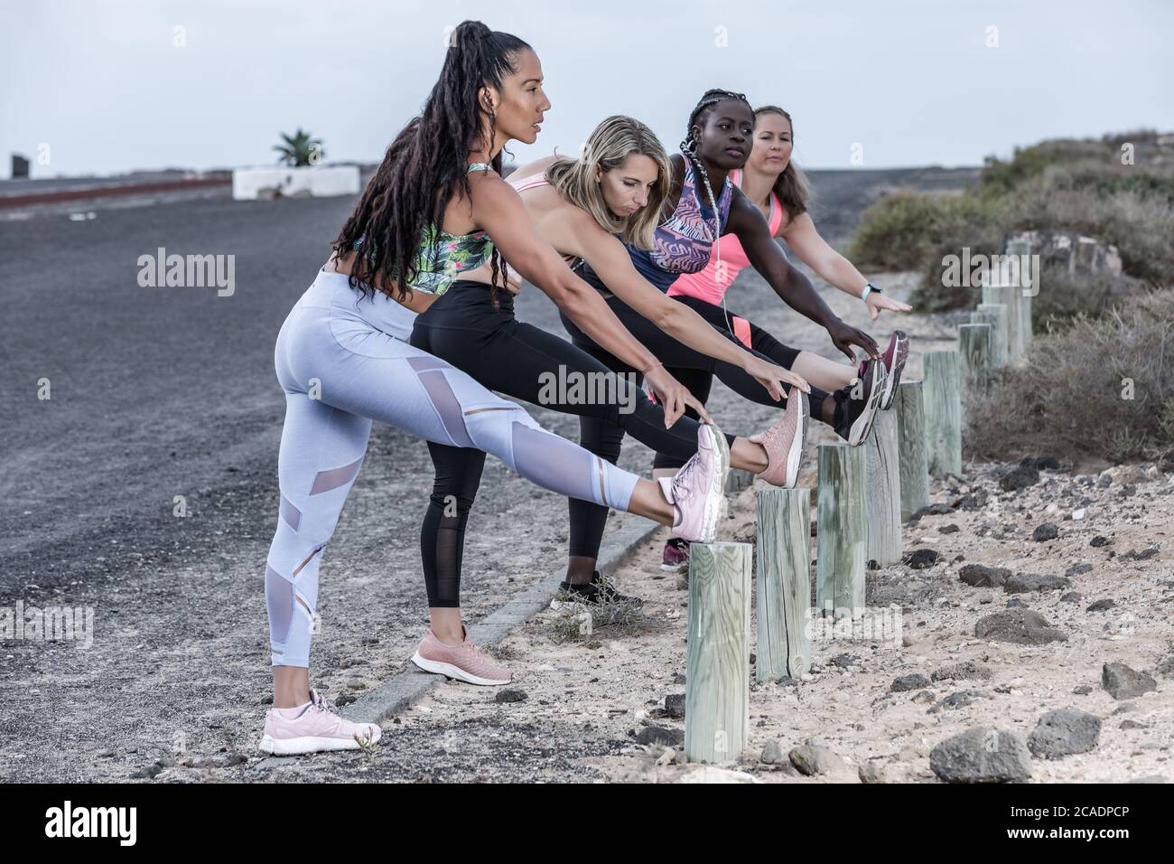 Seitenansicht von vollstämmigen multiethnischen Freundinnen in Sportswear, die die Beine auf Stöcken halten und sich beim Aufwärmen vor dem Laufen nach vorne beugen Stockfoto