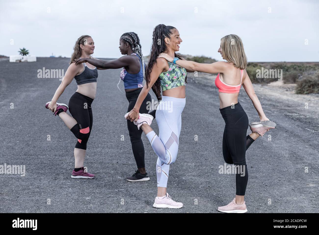 Seitenansicht fröhlicher multirassischer Freundinnen in Sportkleidung, die sich gegenseitig anlehnen und lächeln, während sie gebeugte Beine vor dem Laufen strecken Stockfoto