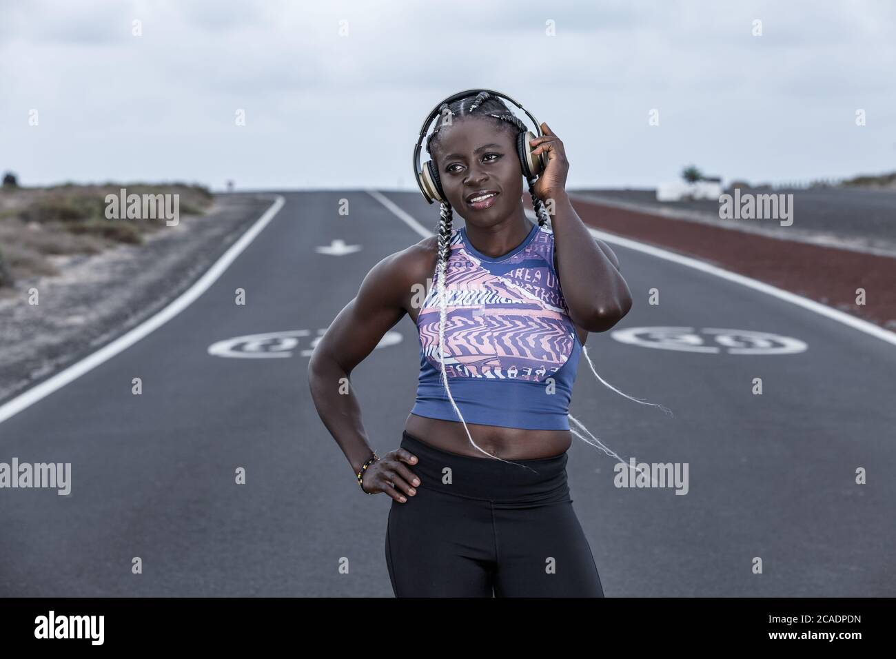 Selbstbewusste afroamerikanische Athletin mit der Hand an der Taille lächelnd und wegblickend, während sie auf der Straße steht und Musik in Kopfhörern duri hört Stockfoto