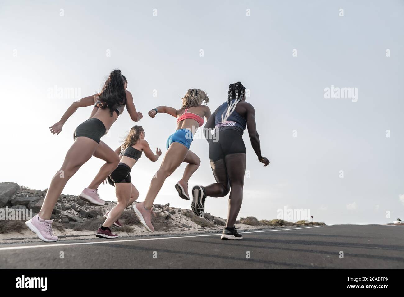 Rückansicht der anonymen multirassischen Sportlerinnen Sprinten auf Asphaltstraße gegen Abendhimmel während Outdoor-Training in Außenbezirken Stockfoto