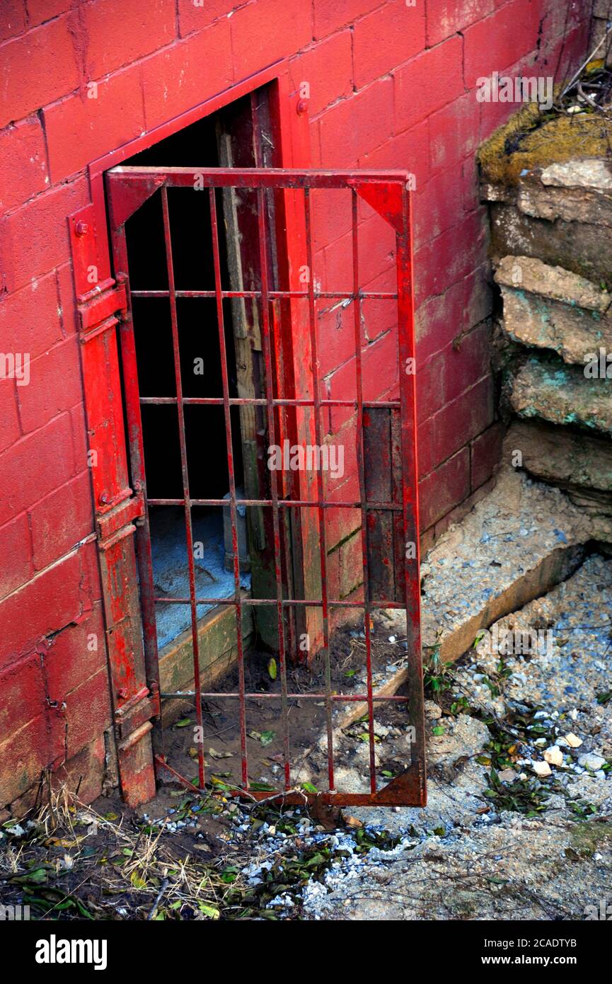 Schmutzige, rostende, rote Metalltür, die offen hing und die Dunkelheit ausließ. Die rote Ziegelwand ist grungig. Stockfoto