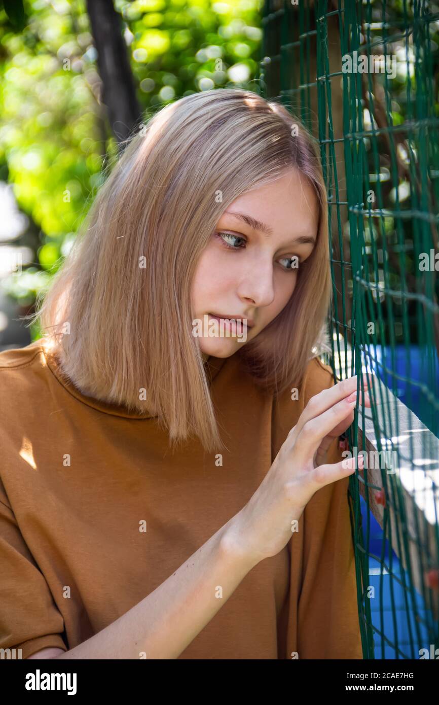 Porträt eines jungen Mädchens, einer Blondine, die sich gegen den Zaun eines Hausgartens lehnt, vor einem Hintergrund aus grünen Blättern Stockfoto