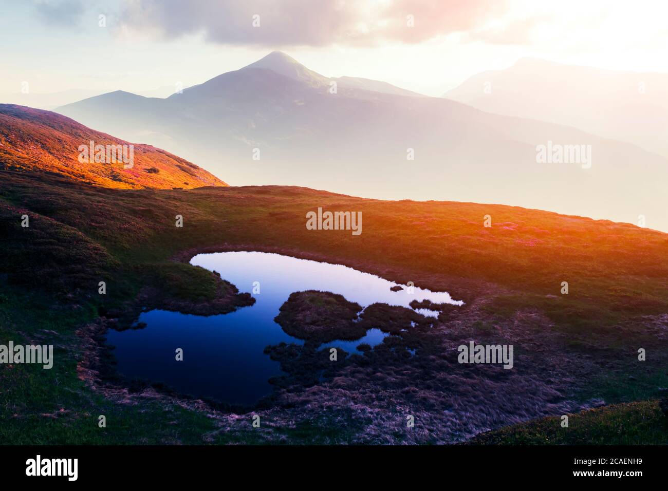 Bergsee bei Sonnenaufgang. Malerische Sommerlandschaft mit grünen Hügeln und Sonnenstrahlen am Morgenhimmel. Karpaten Stockfoto