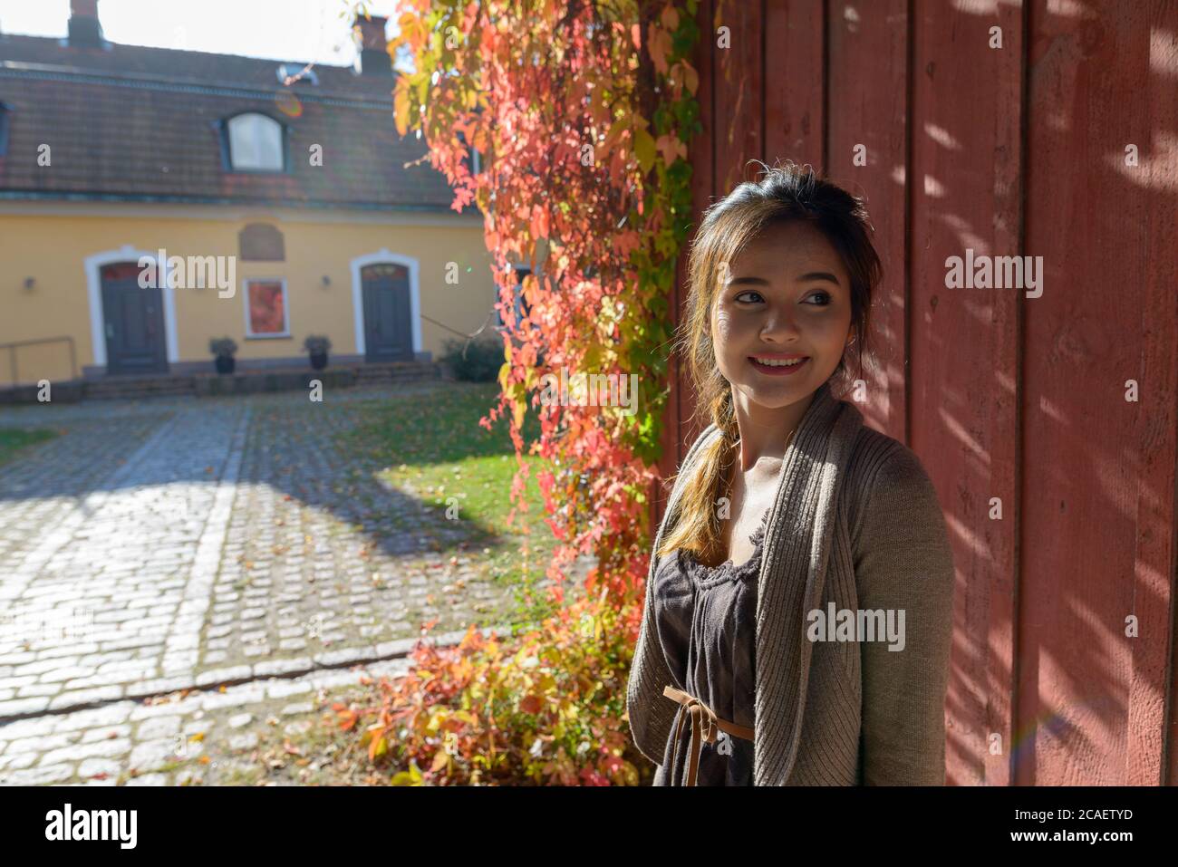 Junge glückliche asiatische Frau lächelt und denkt gegen die Holzwand vor dem eleganten Vorstadthaus Stockfoto