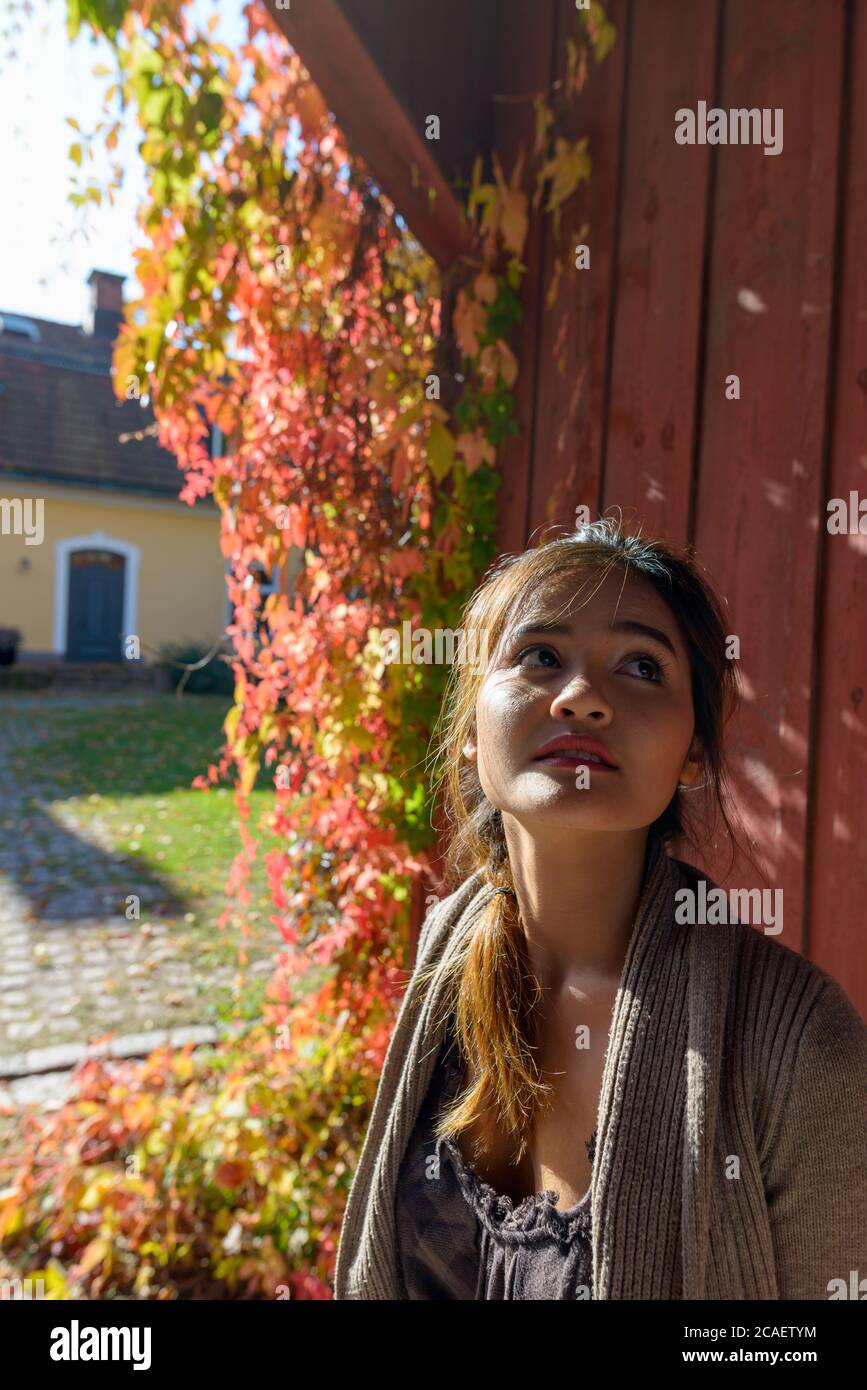 Junge schöne asiatische Frau, die vor einem eleganten Vorstadthaus an einer Holzwand denkt Stockfoto