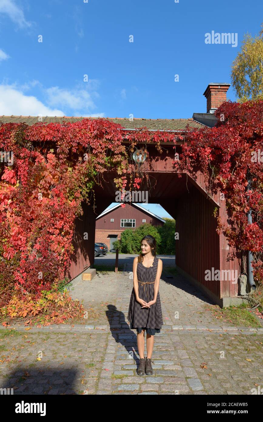 Junge schöne asiatische Frau gegen Vorstadthaus in Herbstblättern bedeckt Stockfoto