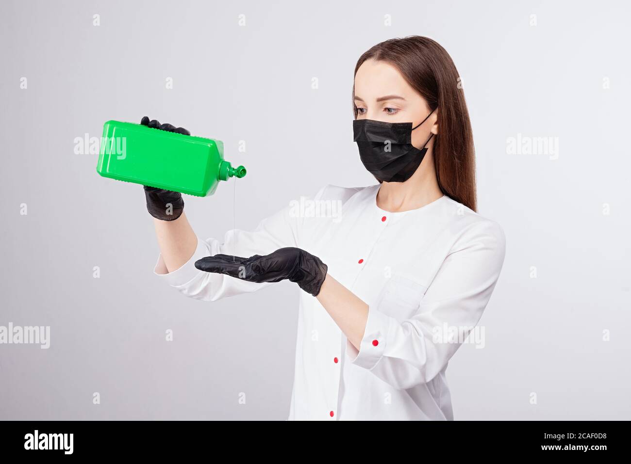 Mädchen in schwarzen Handschuhen und Maske gießt Flüssigkeit aus grünem Glas, Desinfektionslösung, hellgrauer Hintergrund, Frau mit langen Haaren in weißer Kleidung Stockfoto