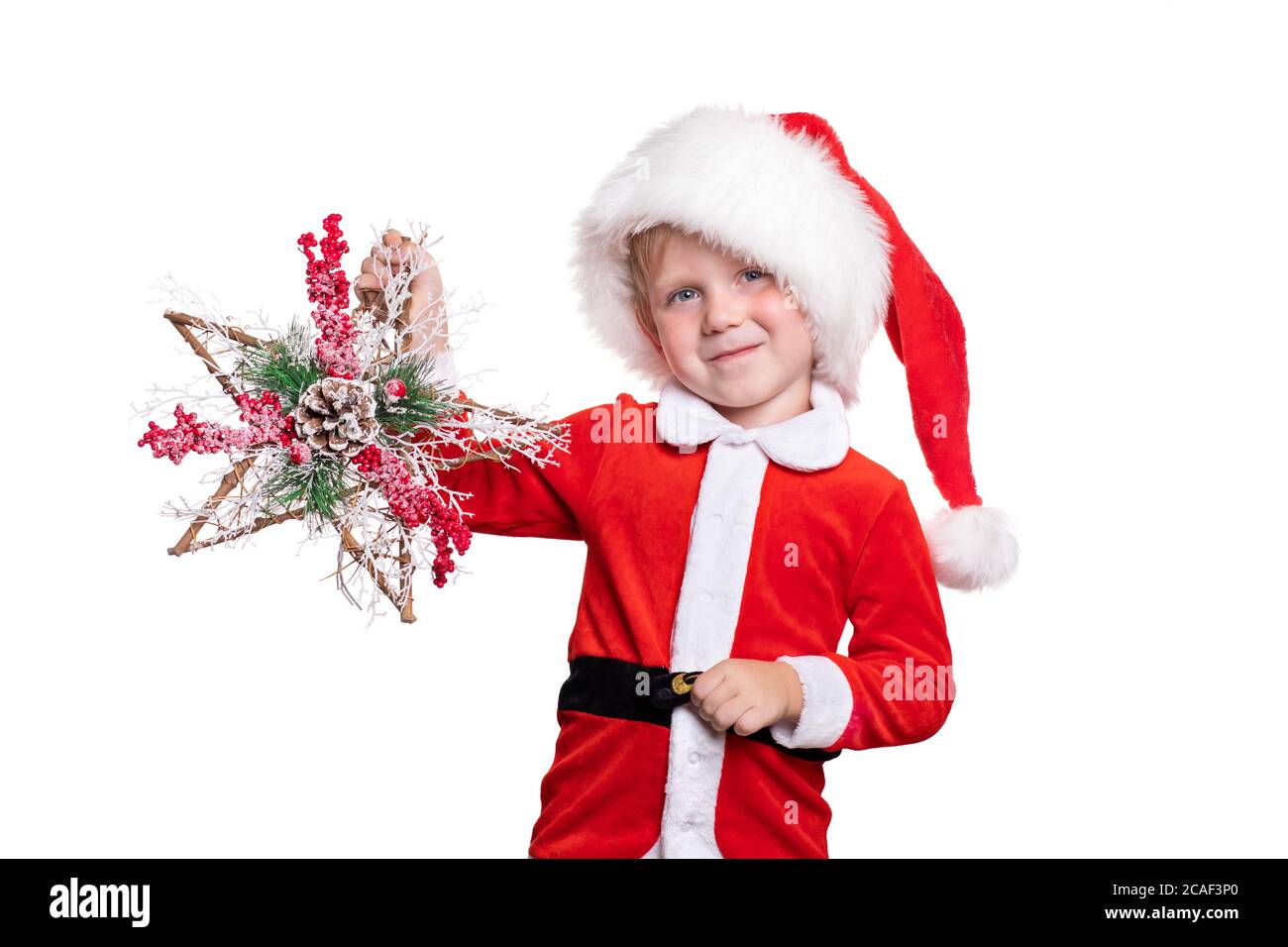 Ein kleiner Junge in einem Anzug und einem Weihnachtsmann Hut hält eine hölzerne Sterndekoration mit Beerenzweigen und einem Pinienzapfen Hände. Neujahrskarte für Weihnachten 20 Stockfoto