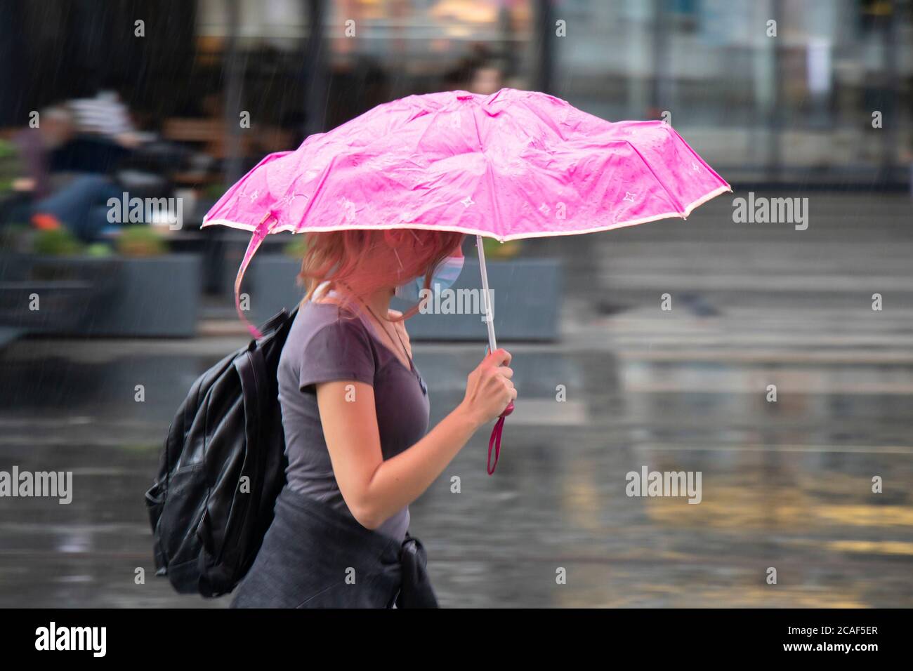 Belgrad, Serbien - 5. August 2020: Bewegungsunschärfe der jungen Frau trägt Gesicht chirurgische Maske zu Fuß schnell unter rosa Regenschirm an einem regnerischen Sommertag in der Stockfoto