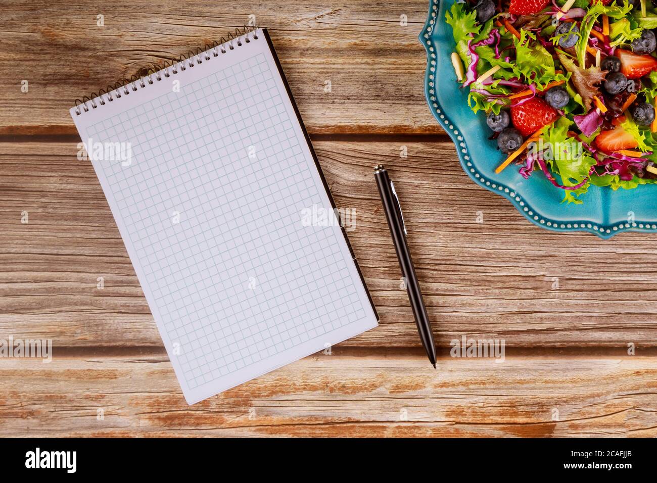 Notizblock und Stift mit Obst- und Gemüsesalat auf Holzhintergrund. Stockfoto