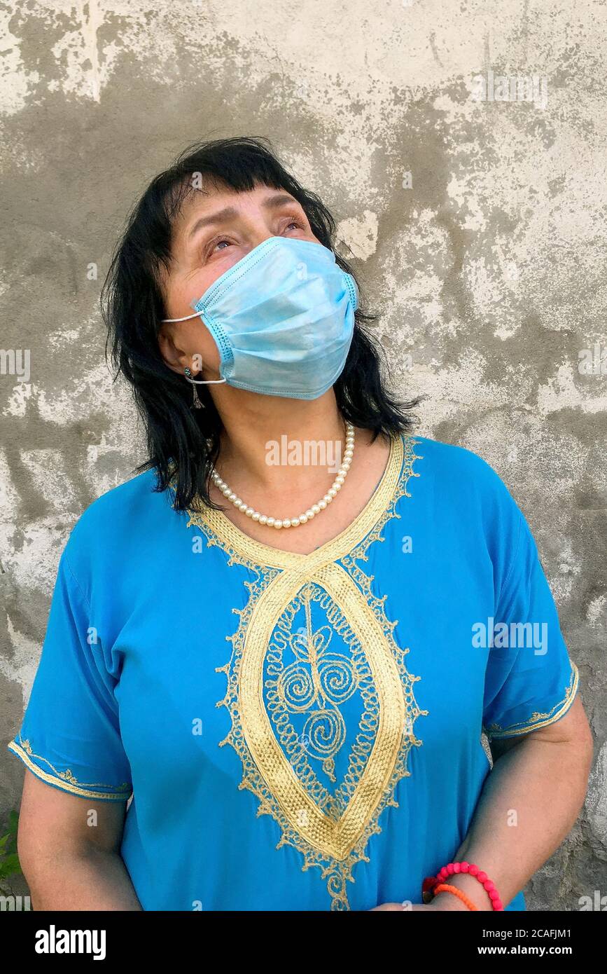 Asiatische oder kaukasische Ethnie reife Frau in blauem Kleid mit medizinischer Maske auf dem Gesicht zu schützen COVID-19, Blick vor Hintergrund von grauen plast Stockfoto