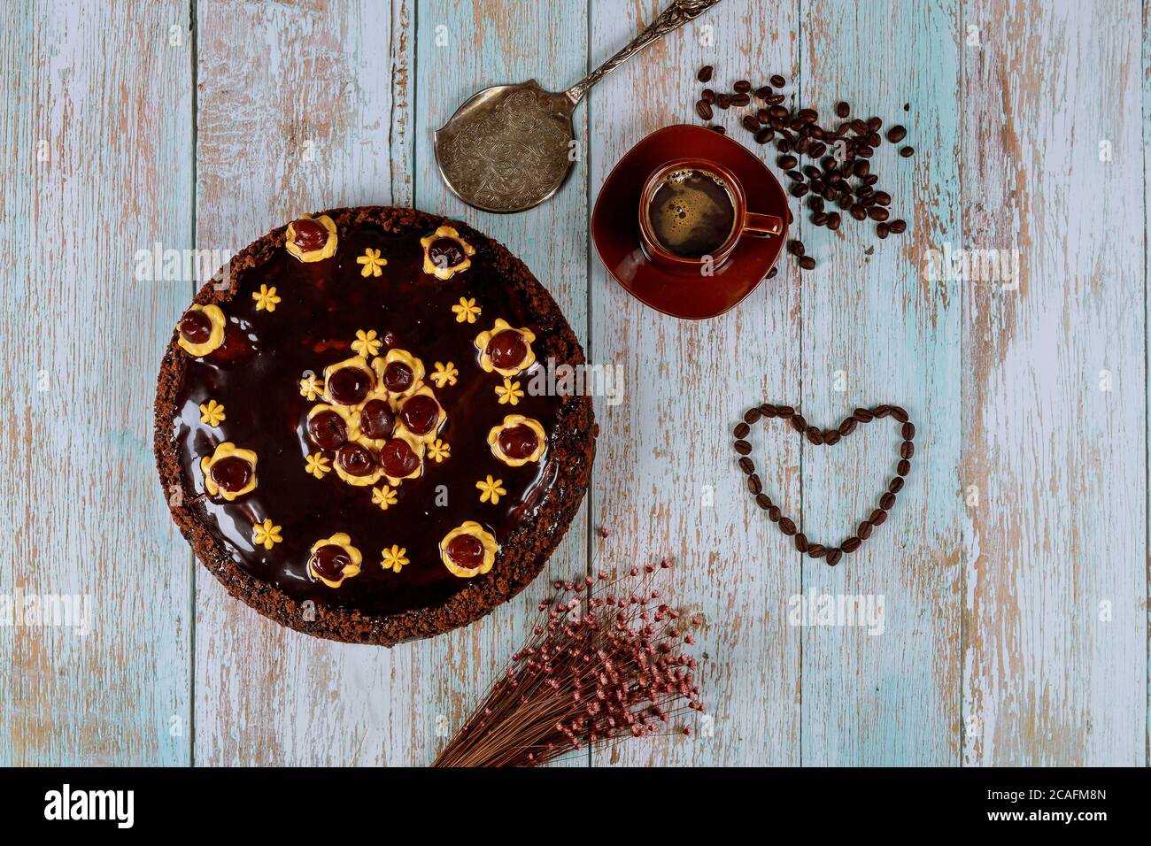 Kirschkuchen mit Schokoladenglasur, Tasse Kaffee und Blumen auf Holzgrund. Stockfoto
