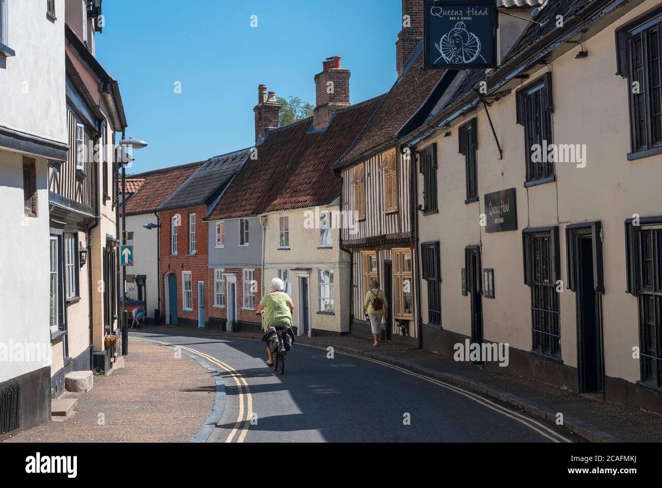 Norfolk Sommer Großbritannien, Rückansicht im Sommer einer älteren Frau, die entlang einer typischen Marktstadtstraße in Wymondham, Norfolk, England, Großbritannien radelt Stockfoto