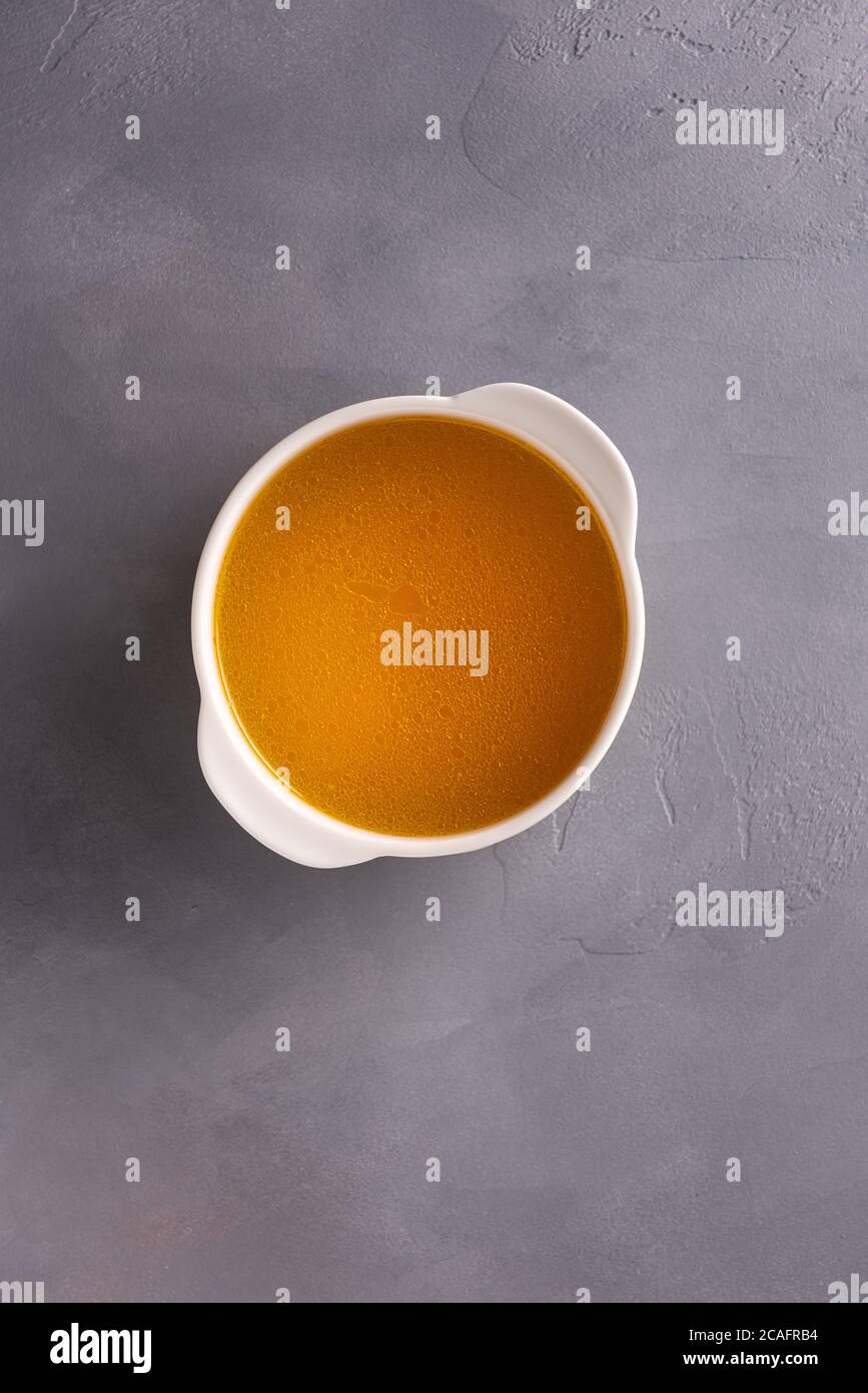 Hühnerbouillon mit dünner Fettschicht auf der Oberfläche in kleiner weißer Suppenschüssel auf strukturierter grauer Oberfläche. Direkt über der Ansicht. Vertikaler Hintergrund mit la Stockfoto