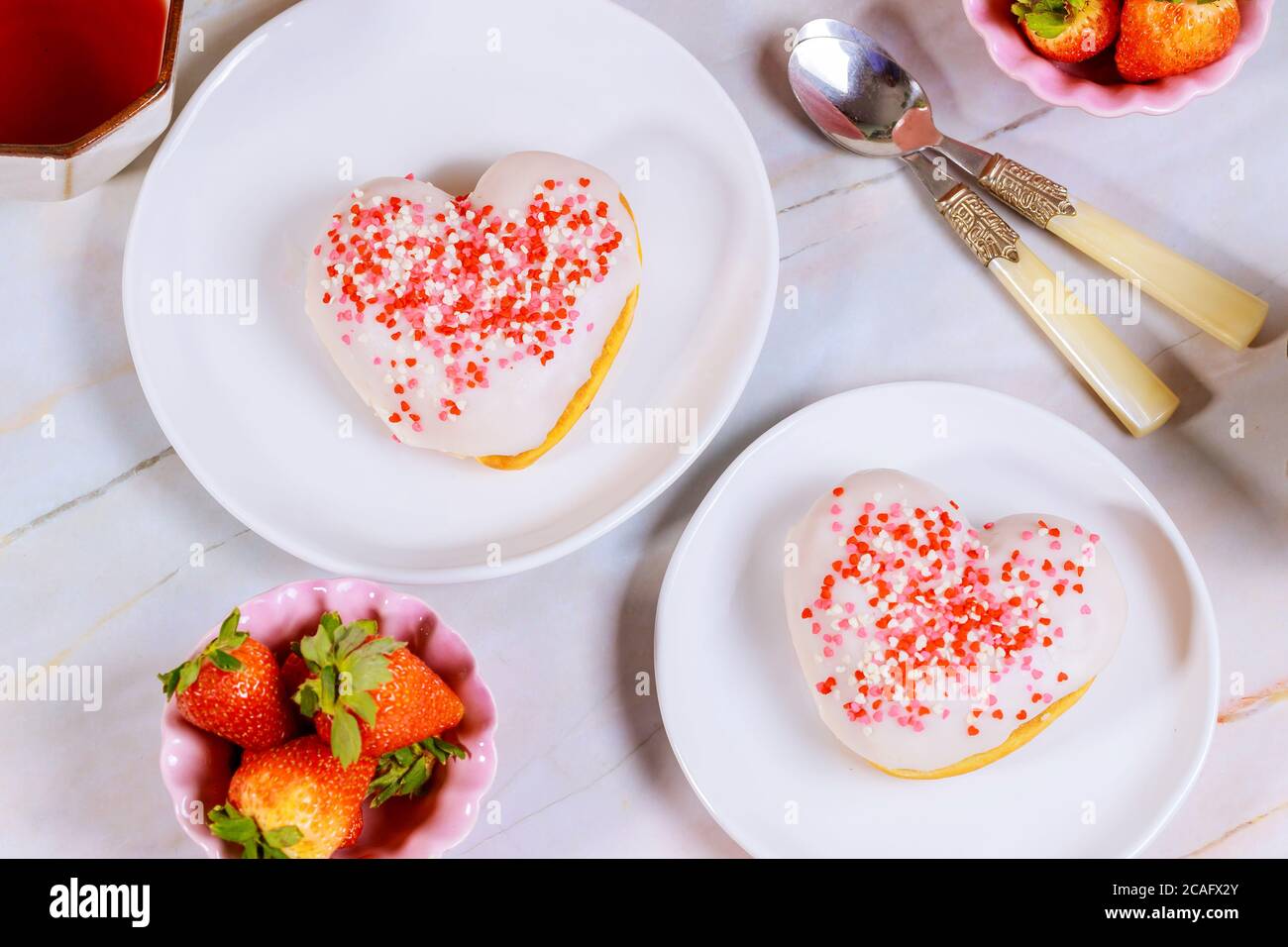Zwei herzförmige Donuts mit weißer Glasur und rosa, roten Streuseln auf einer Platte mit Erdbeere und Löffel. Valentinstag-Konzept. Stockfoto