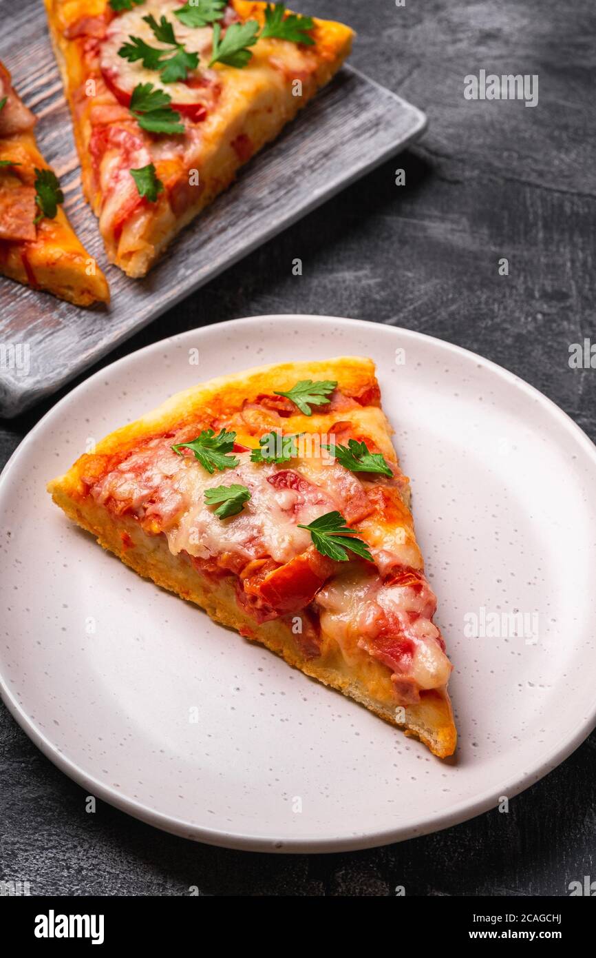 Heiße Pizzascheiben mit Mozzarella-Käse, Schinken, Tomate und Petersilie auf Holz Schneidebrett und Teller, Stein Beton Hintergrund, Ansicht Makro Stockfoto