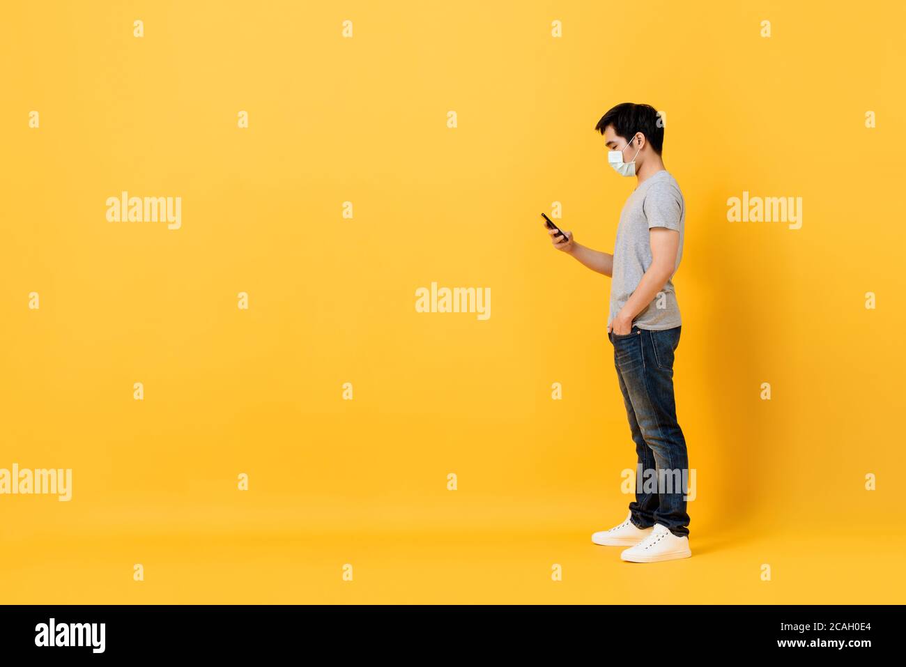 Junge asiatische Mann trägt medizinische Gesichtsmaske stehen und mit Mobiltelefon isoliert auf gelbem Studiohintergrund mit Kopierplatz Stockfoto