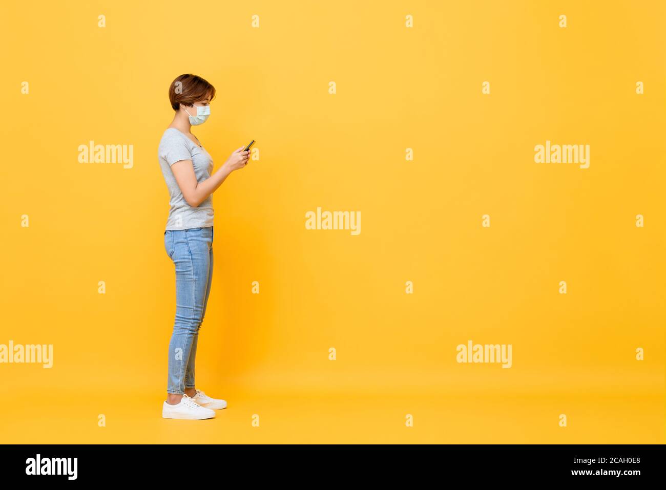 Junge asiatische Frau trägt medizinische Gesichtsmaske stehen und mit Mobiltelefon isoliert auf gelbem Studiohintergrund mit Kopierplatz Stockfoto