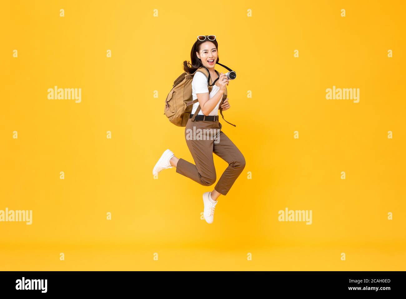 Junge hübsche asiatische Frau Tourist Backpacker lächelnd und springen mit Kamera in der Hand isoliert auf gelbem Hintergrund Stockfoto