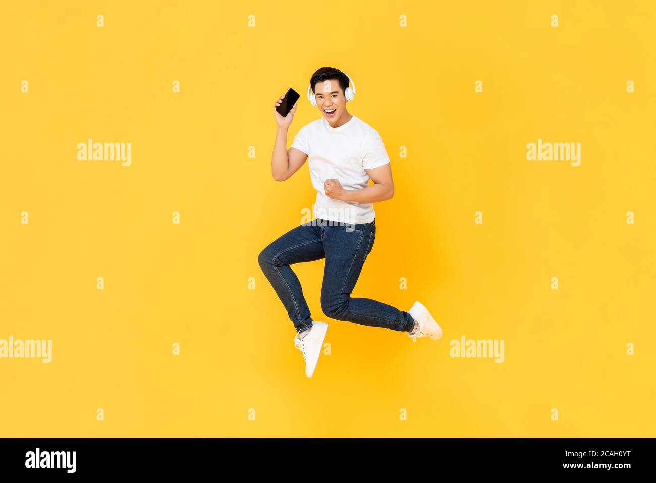 Glücklich junge asiatische Mann trägt Kopfhörer hören Musik aus Handy und springen isoliert auf gelbem Hintergrund Stockfoto