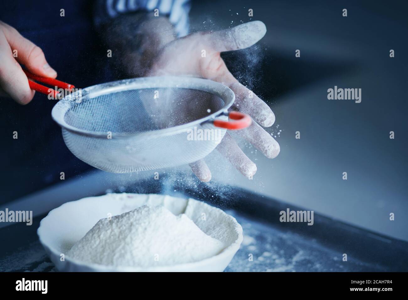 Ein Koch in einem gestreiften Hemd siebt Weizenmehl durch ein großes Sieb in eine weiße Schüssel und verschmutzt seine Hände mit Mehl. Der Prozess der Vorbereitung Teig für BA Stockfoto