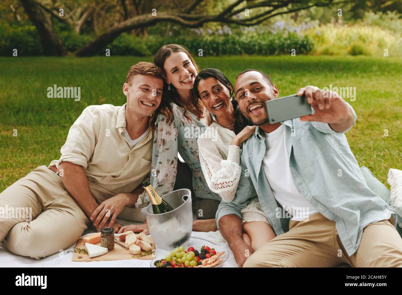 Junger Mann, der beim Picknick Selfie mit Freunden nimmt. Multiethnische Gruppe junger Männer und Frauen, die für ein Selfie im Park posieren. Stockfoto
