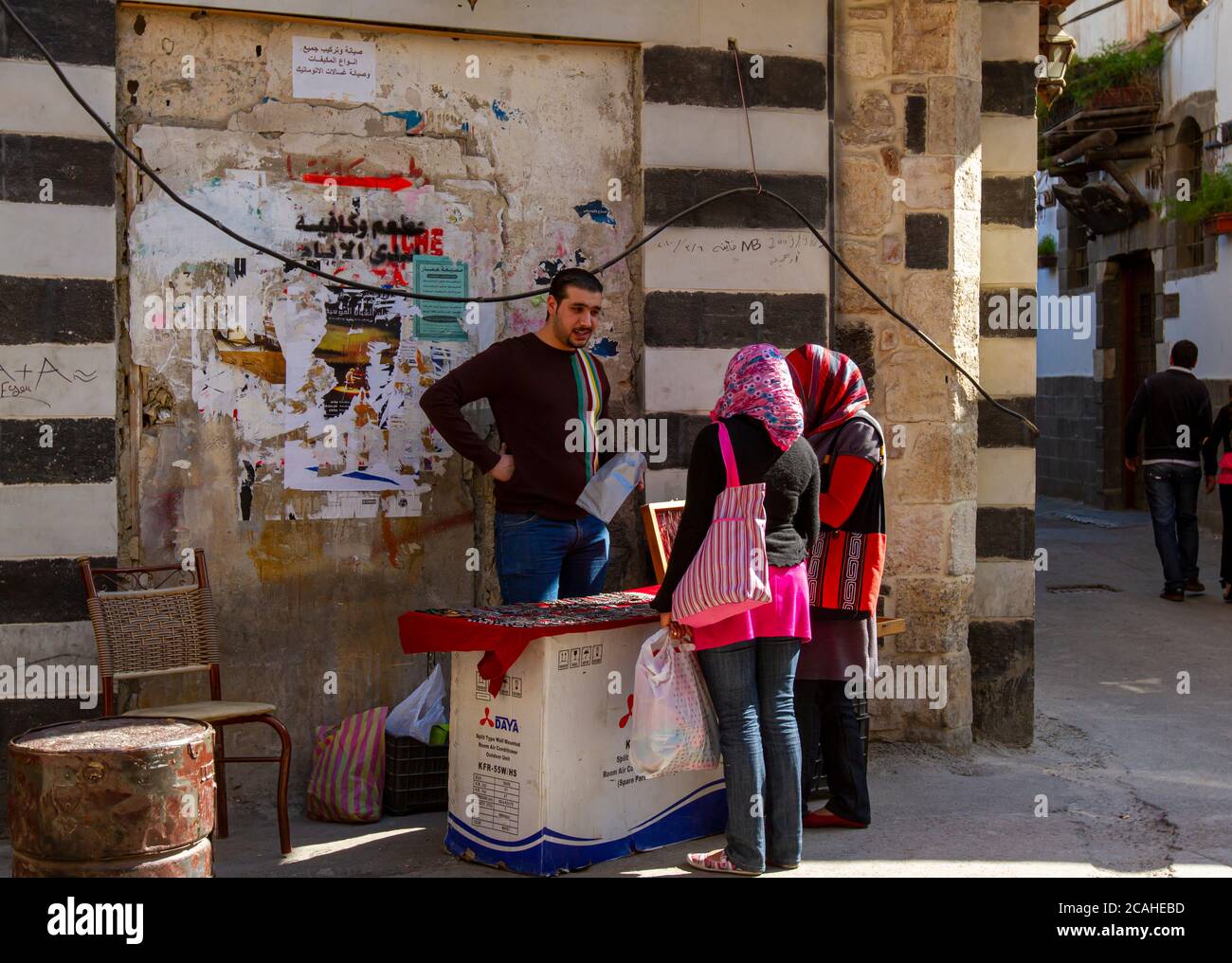 Damaskus, Syrien 03/29/2010: Ein junger, gut aussehender Verkäufer der syrischen Straße versucht, Ornamente an zwei arabische Frauen zu verkaufen. Er zeigt Elemente auf Notbehelfsbehelf an Stockfoto