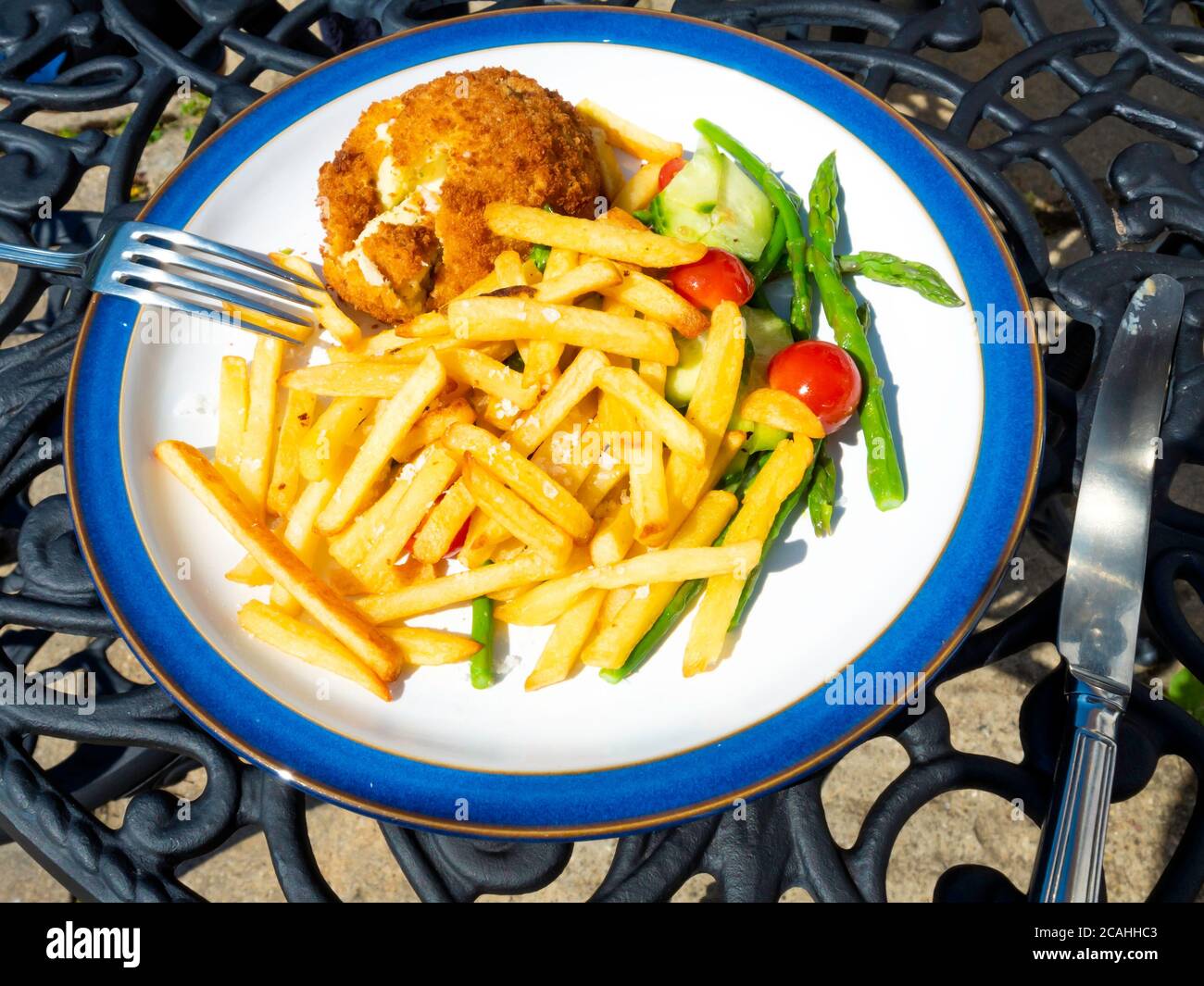 Ein Mittagessen mit geräuchertem Schellfisch-Kuchen mit französischen Bratkartoffeln, Spargeltomaten und Gurken, serviert im Freien auf einem Außentisch Stockfoto