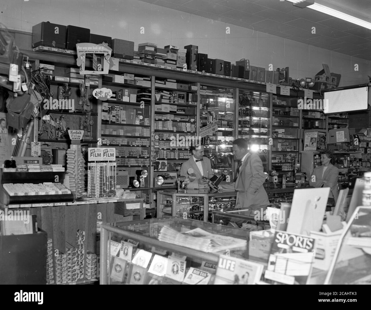 1940er Jahre, historisch, an der Theke, in einem großen Fotoladen, ein elegant gekleideter Verkaufsvertreter mit Fliege, der einem männlichen Kunden eine der neuesten Mittelformat-Filmkameras mit Faltenlinse oder Faltenbalg zeigt, USA. Stockfoto