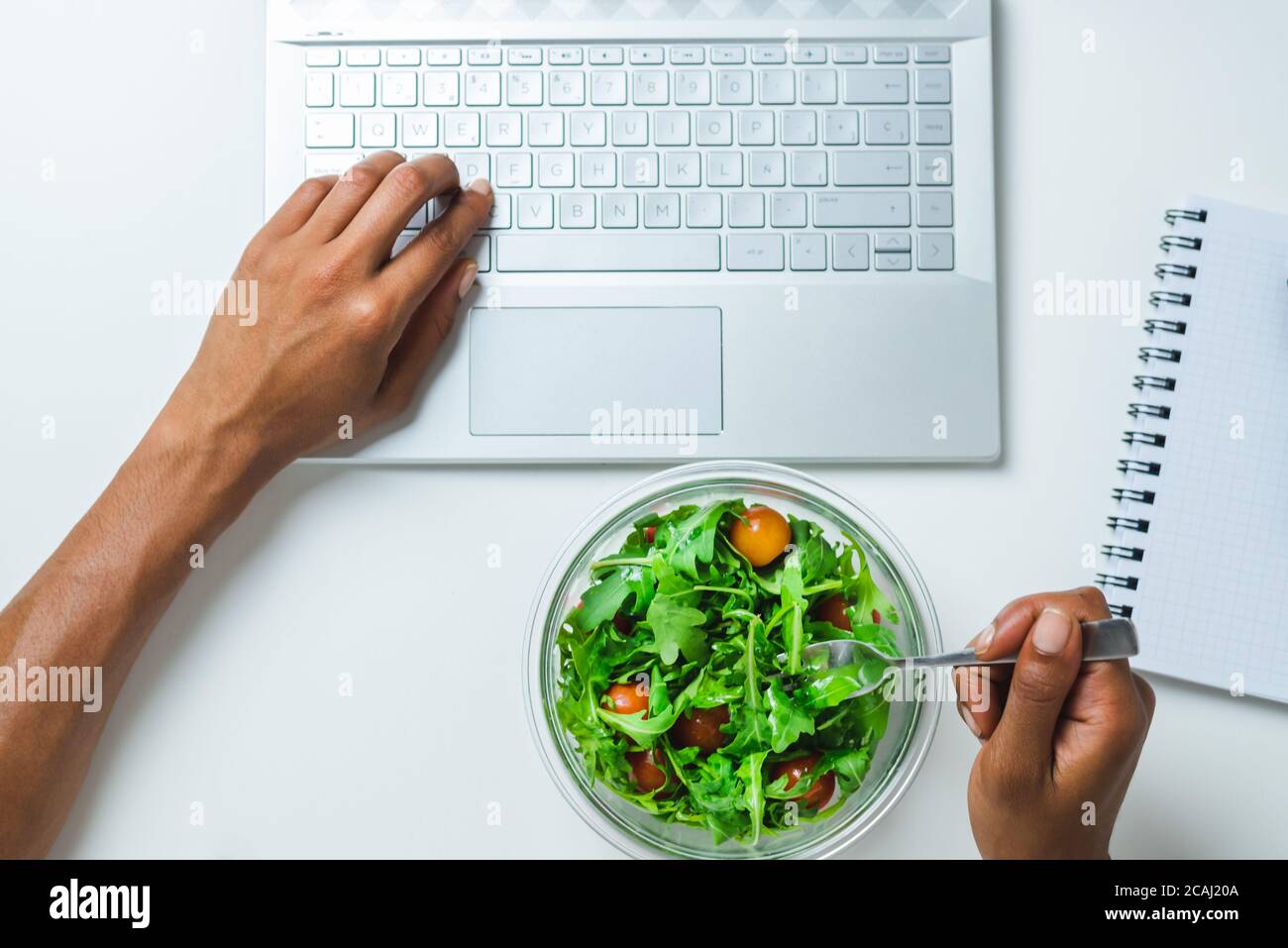 Draufsicht auf die Hände einer schwarzen Frau mit Ein Laptop beim Essen einen Salat auf der Oberseite Ein weißer Schreibtisch Stockfoto